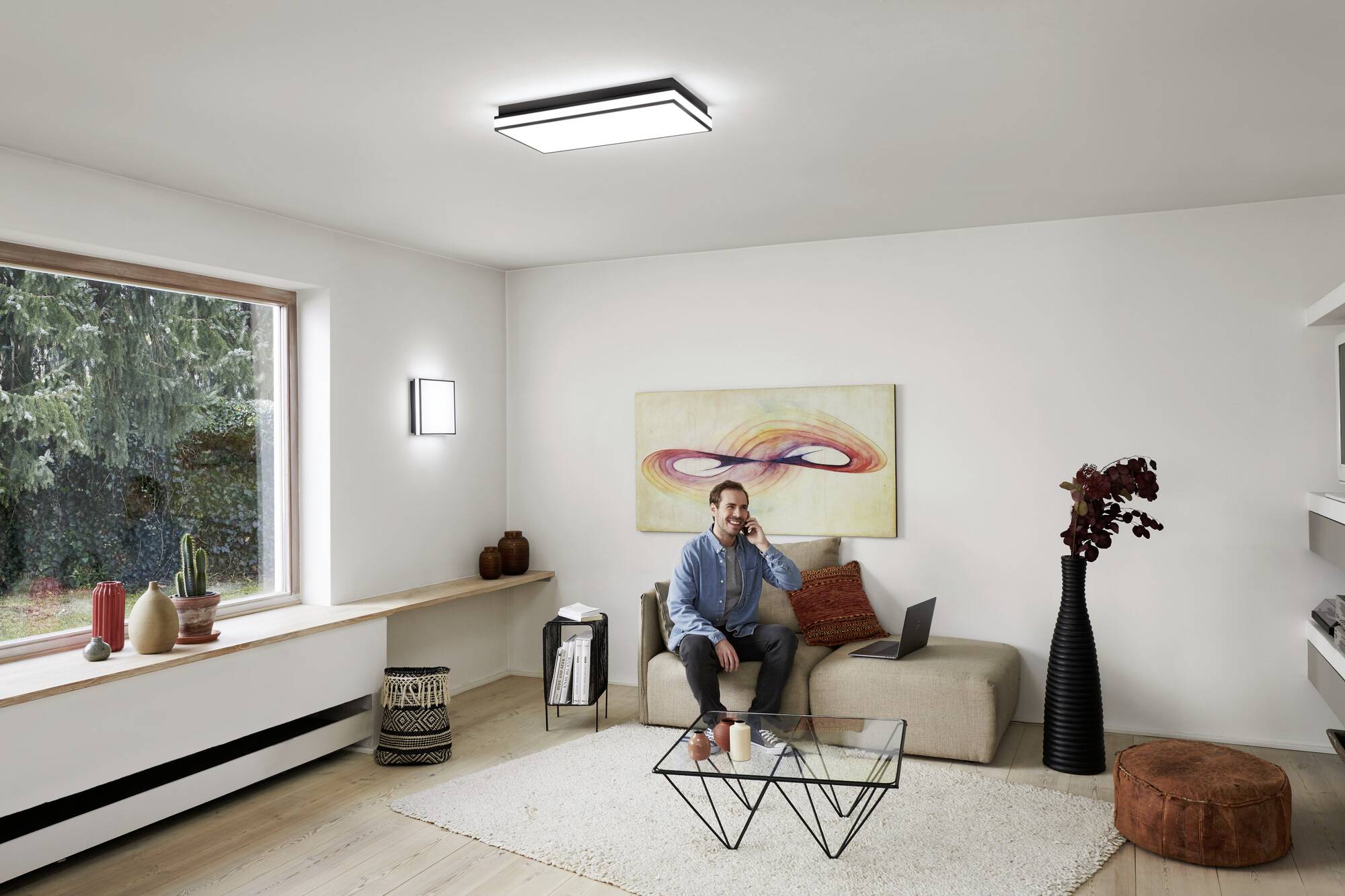 Ein Mann sitzt auf einem Sofa in einem modernen Wohnzimmer, führt ein Telefonat und hat einen Laptop vor sich. Ein großes Fenster bietet Blick nach draußen.