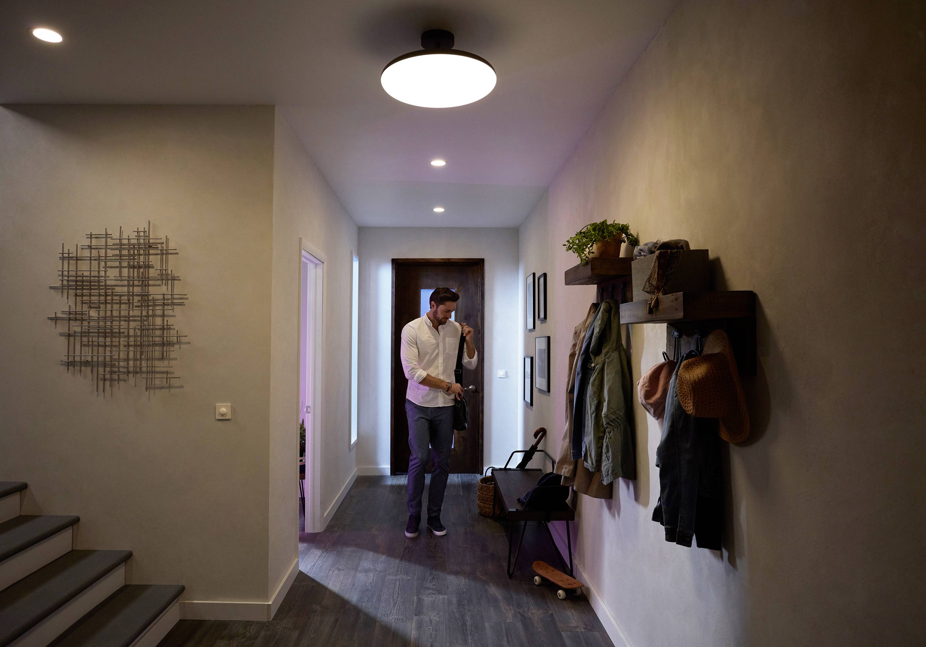 Ein Mann im Flur zieht eine Jacke an. An der Wand hängen Mäntel und Hüte, Regale mit Pflanzen sind sichtbar. Warmes Licht beleuchtet den Raum.
