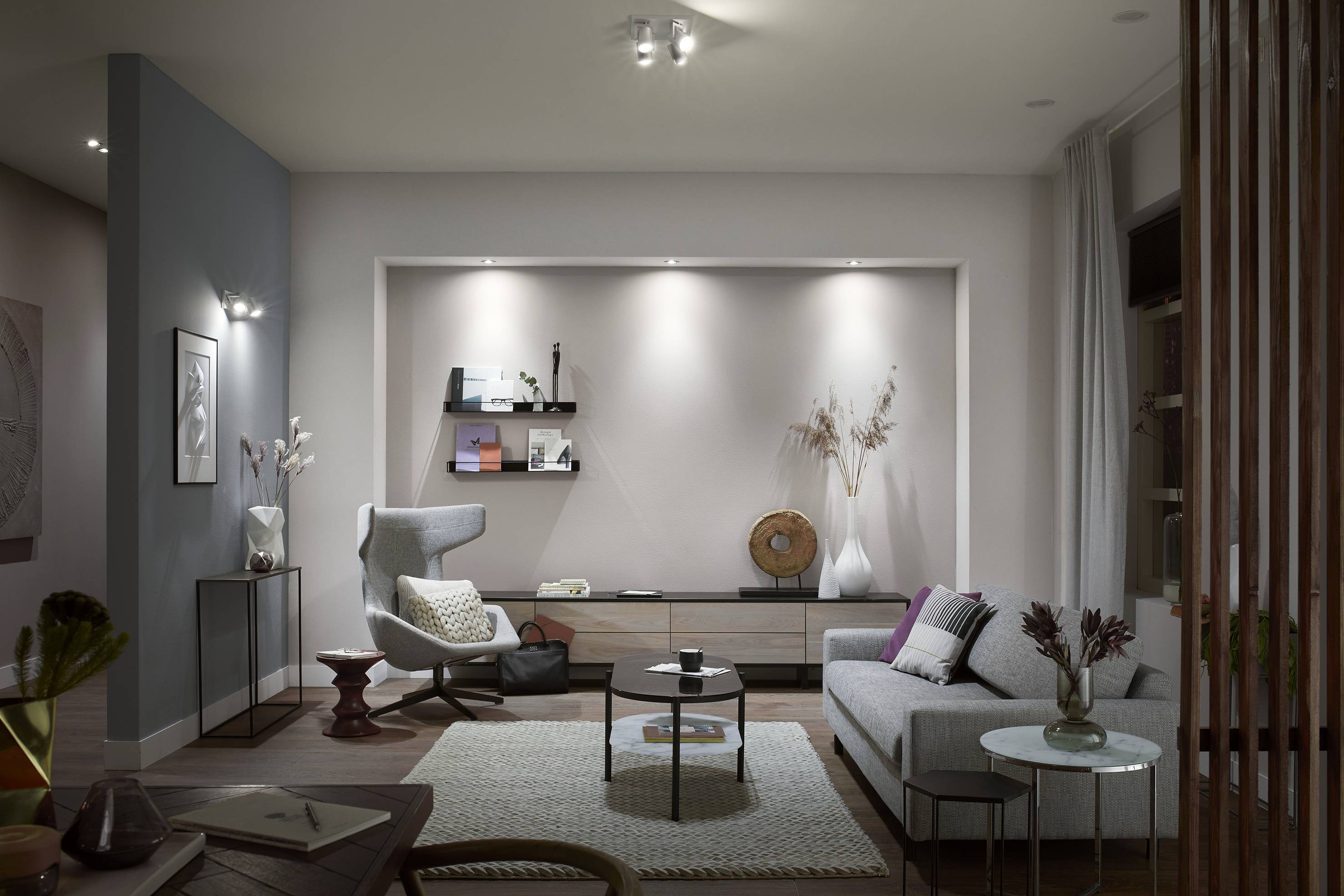 Ein modernes Wohnzimmer mit einem grauen Sofa, einem Sessel, einem runden Couchtisch und dekorativen Regalen an der Wand.