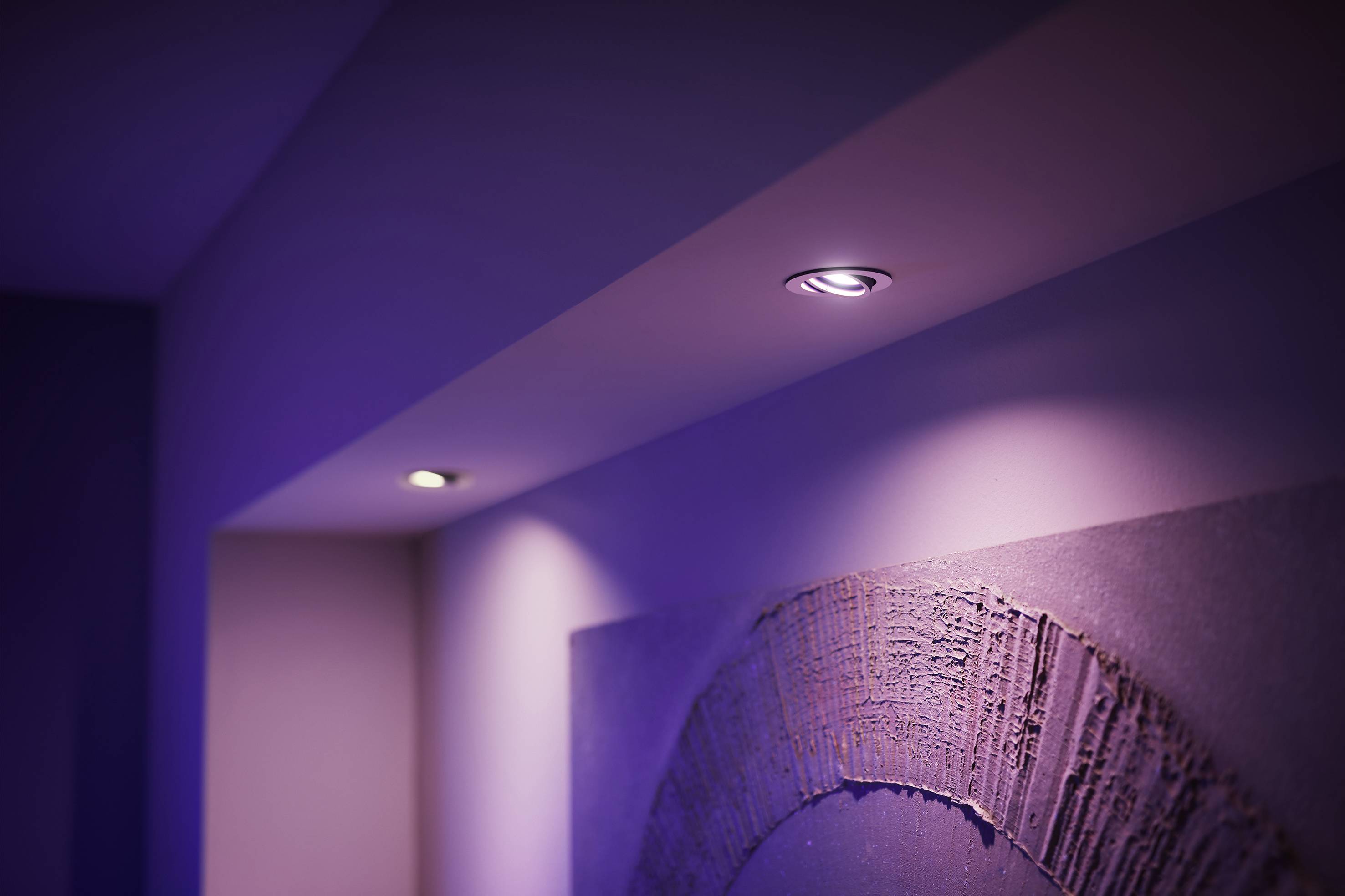 Eine Decke mit eingelassenen LED-Lichtern beleuchtet ein abstraktes Kunstwerk an der Wand, das in violettem Licht erscheint.