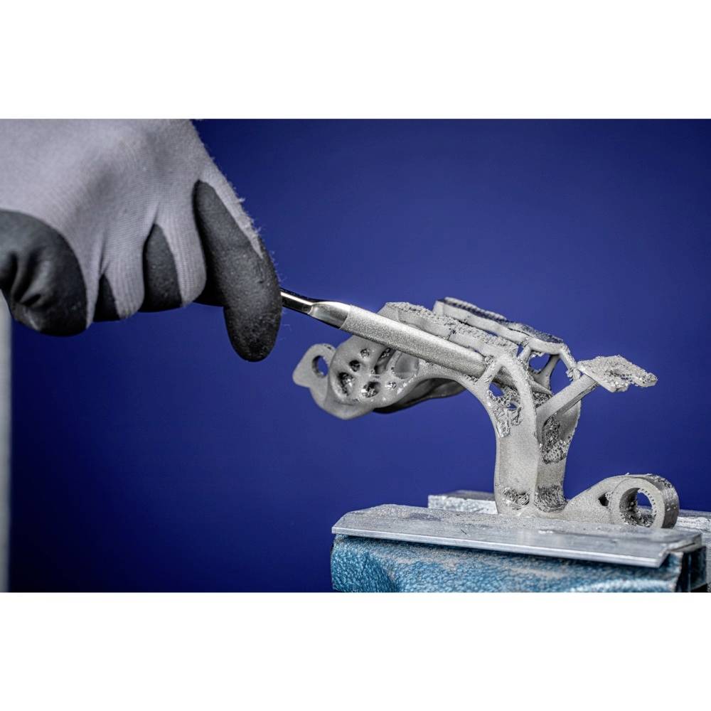 Ein Handschuh hält ein Werkzeug und bearbeitet ein komplexes Bauteil aus einem 3D-Drucker auf einer Werkbank vor blauem Hintergrund.