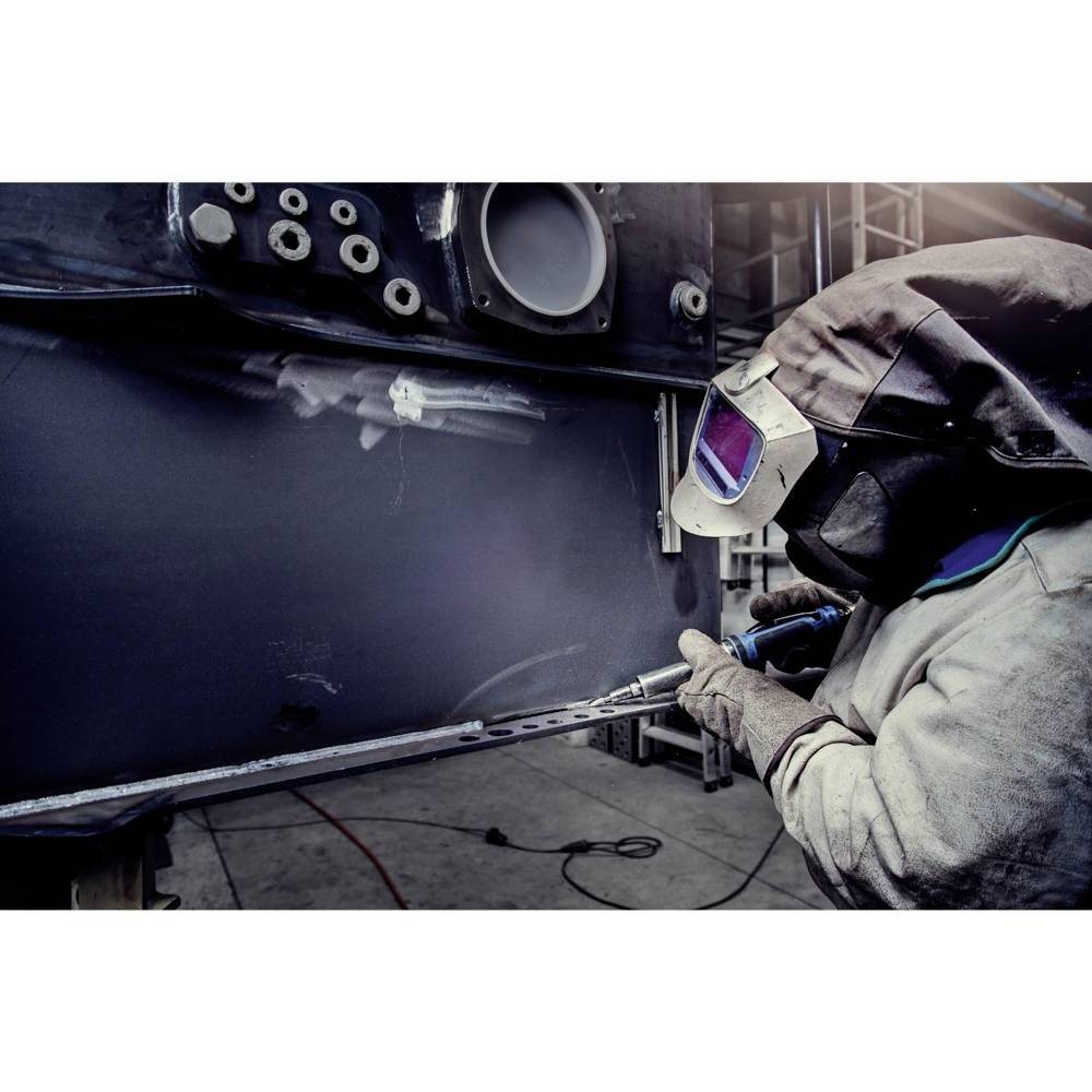 Ein Schweißer im Schutzanzug und Helm arbeitet mit einem Schweißgerät an einem großen Metallstück in einer Fabrikumgebung.