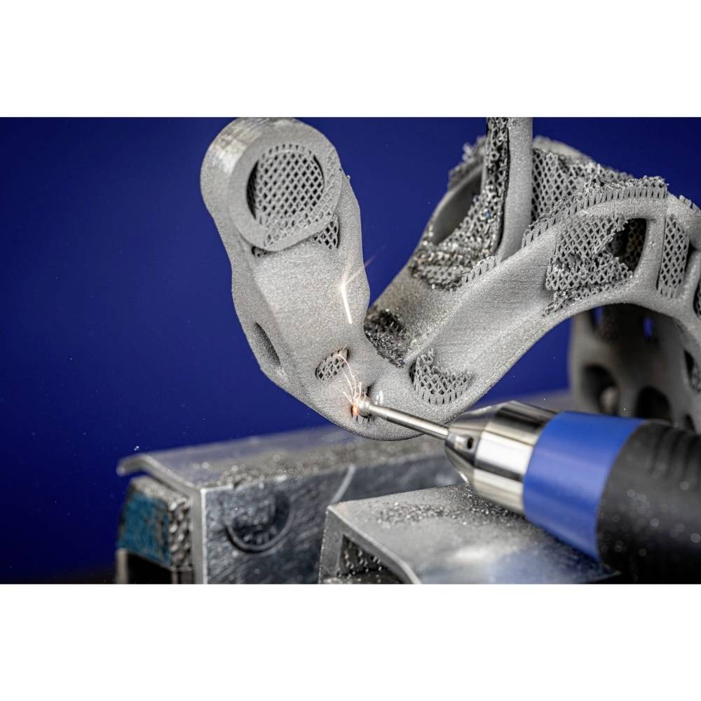 'Nahaufnahme einer 3D-Druckkomponente, die mit einem blauen Werkzeug bearbeitet wird. Funken sprühen bei der Bearbeitung des Metallstücks.'<br><br>
