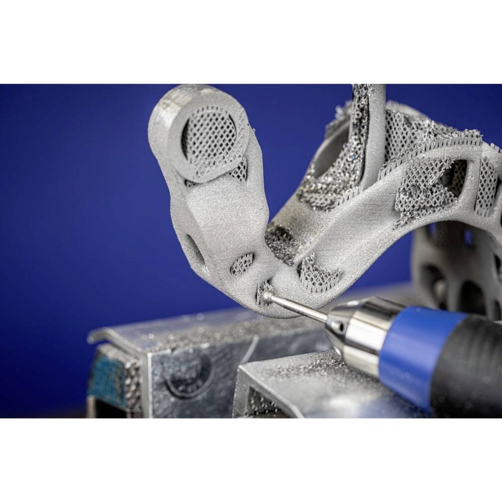 Nahaufnahme eines 3D-gedruckten Metallteils, das mit einem blauen Werkzeug bearbeitet wird. Das Teil zeigt komplexe Strukturen und Details.