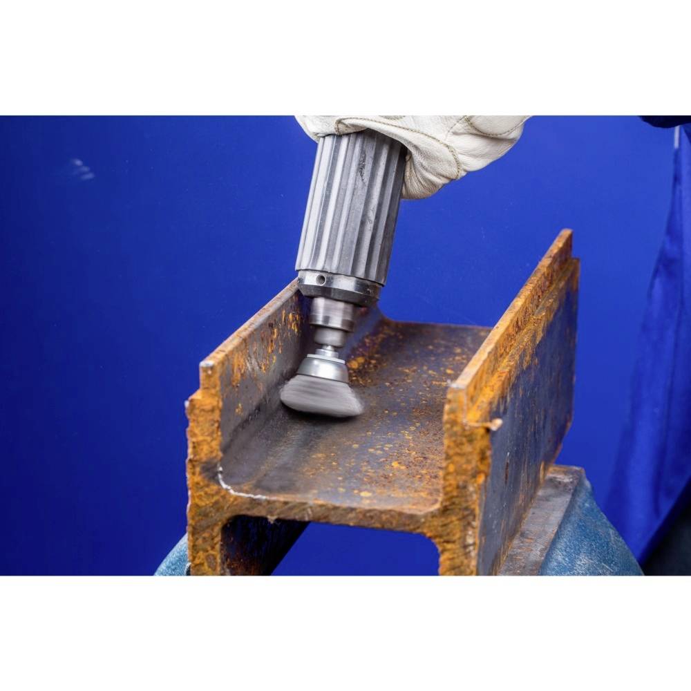 Eine Hand mit Handschuh hält ein Schleifwerkzeug und bearbeitet die verrostete Innenseite eines Stahlträgers vor einem blauen Hintergrund.