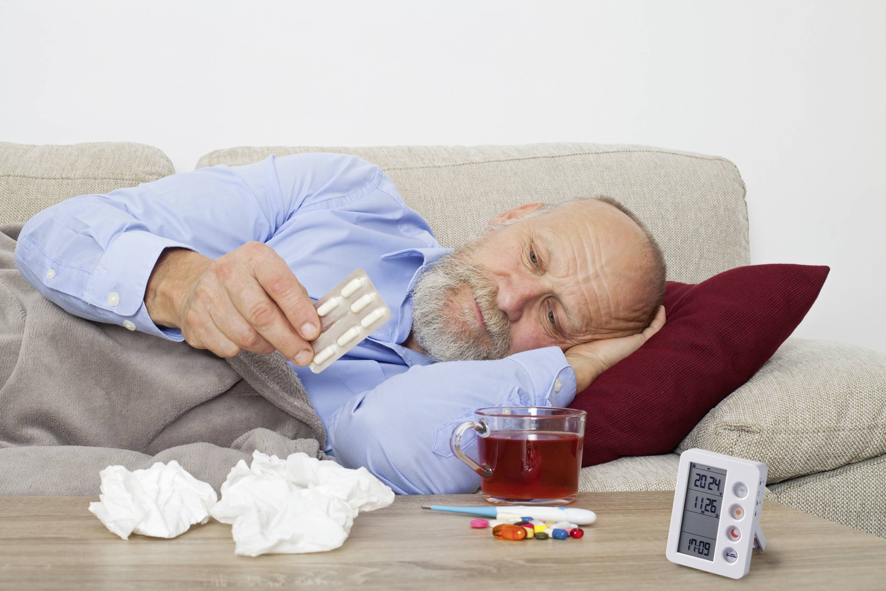 Ein älterer Mann liegt krank auf dem Sofa, hält Medikamente in der Hand, umgeben von Taschentüchern, Tee und einem Thermometer.