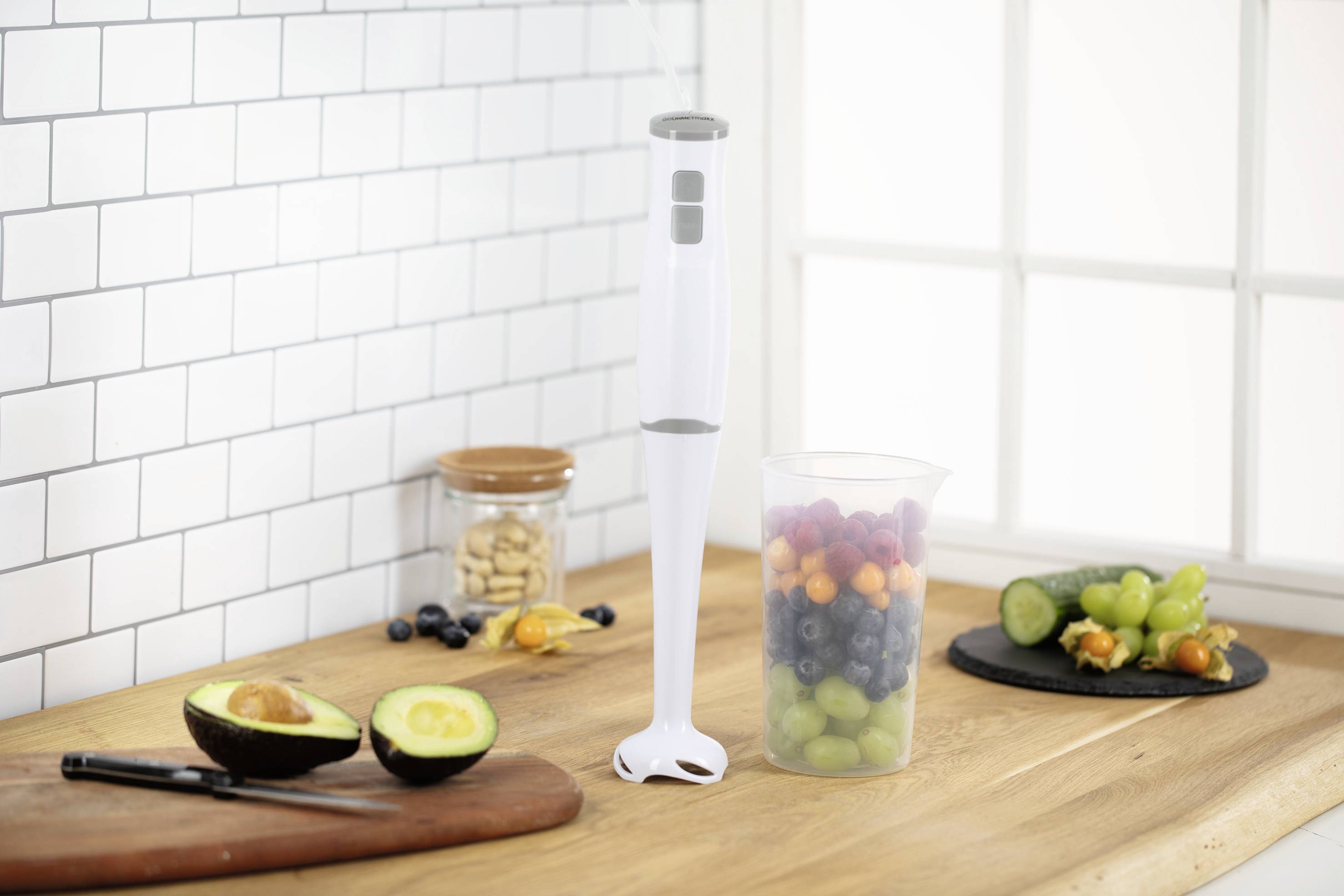 Ein Stabmixer steht auf einer Küchenarbeitsplatte neben einem aufgeschnittenen Avocado, einem Glas Nüsse und einem Becher voller Beeren.