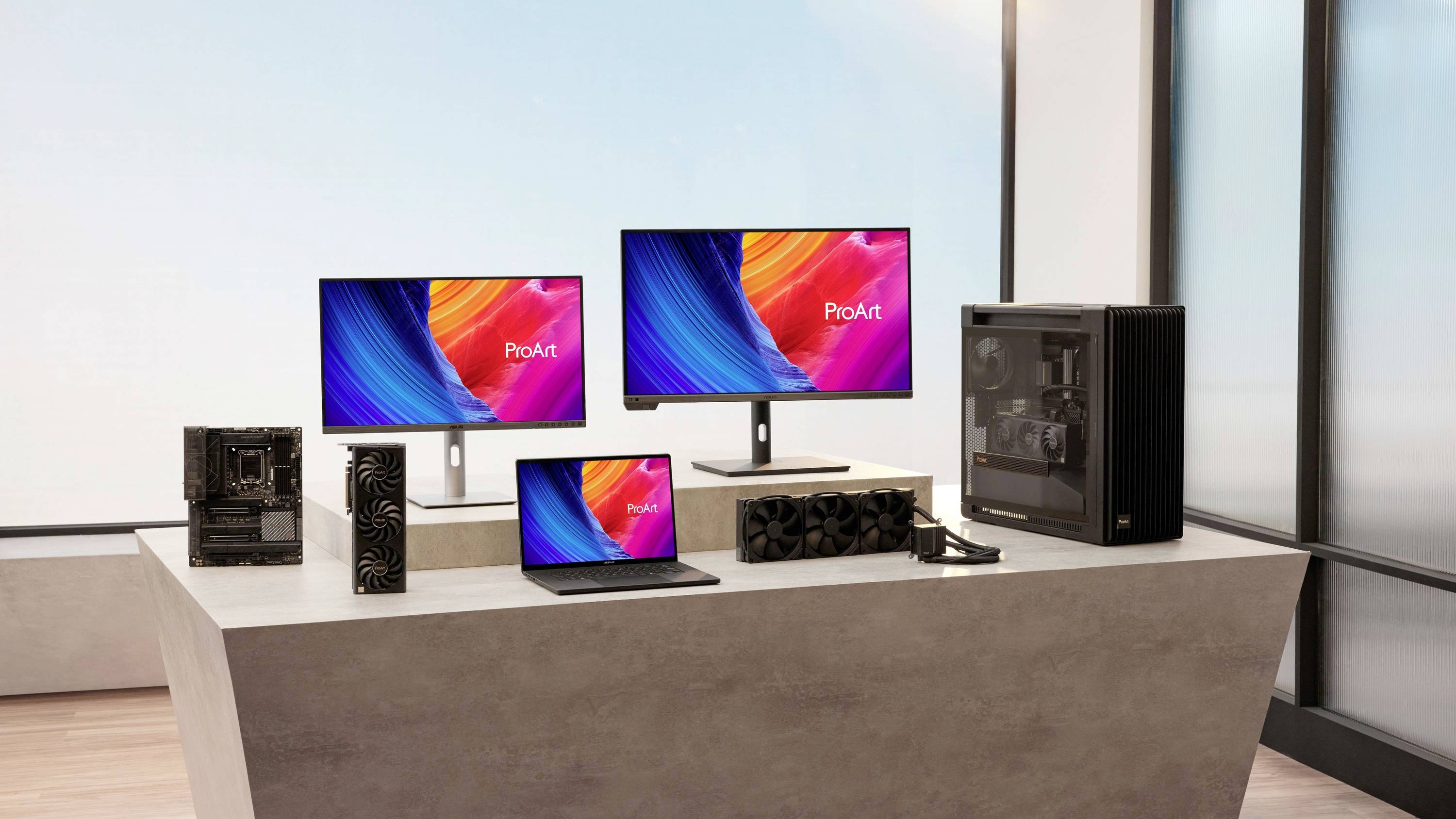 Mehrere Computergeräte auf einem Tisch: zwei Monitore mit 'ProArt'-Bildschirm, ein Laptop, ein Desktop-PC, eine Grafikkarte und ein Mainboard.