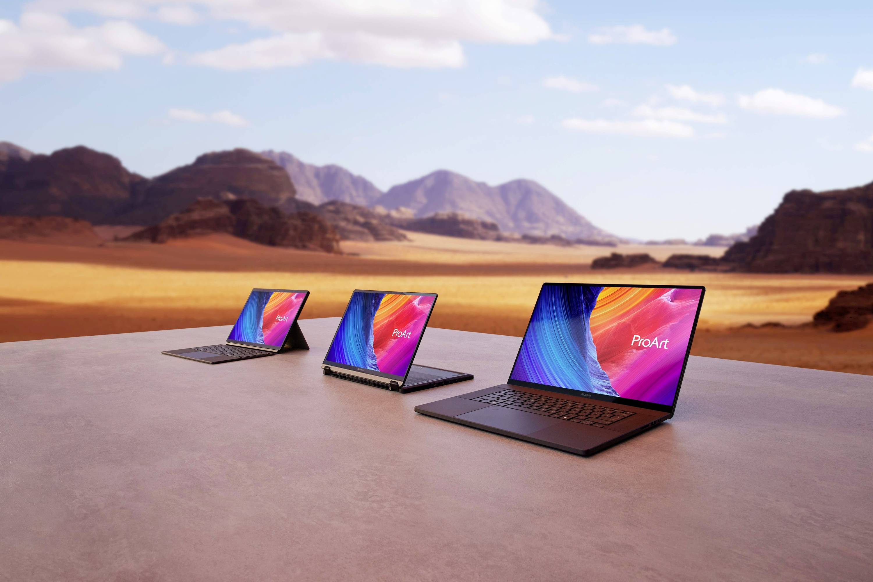 Drei Laptops mit bunten Bildschirmen stehen auf einem Tisch in einer Wüstenlandschaft mit Bergen im Hintergrund.