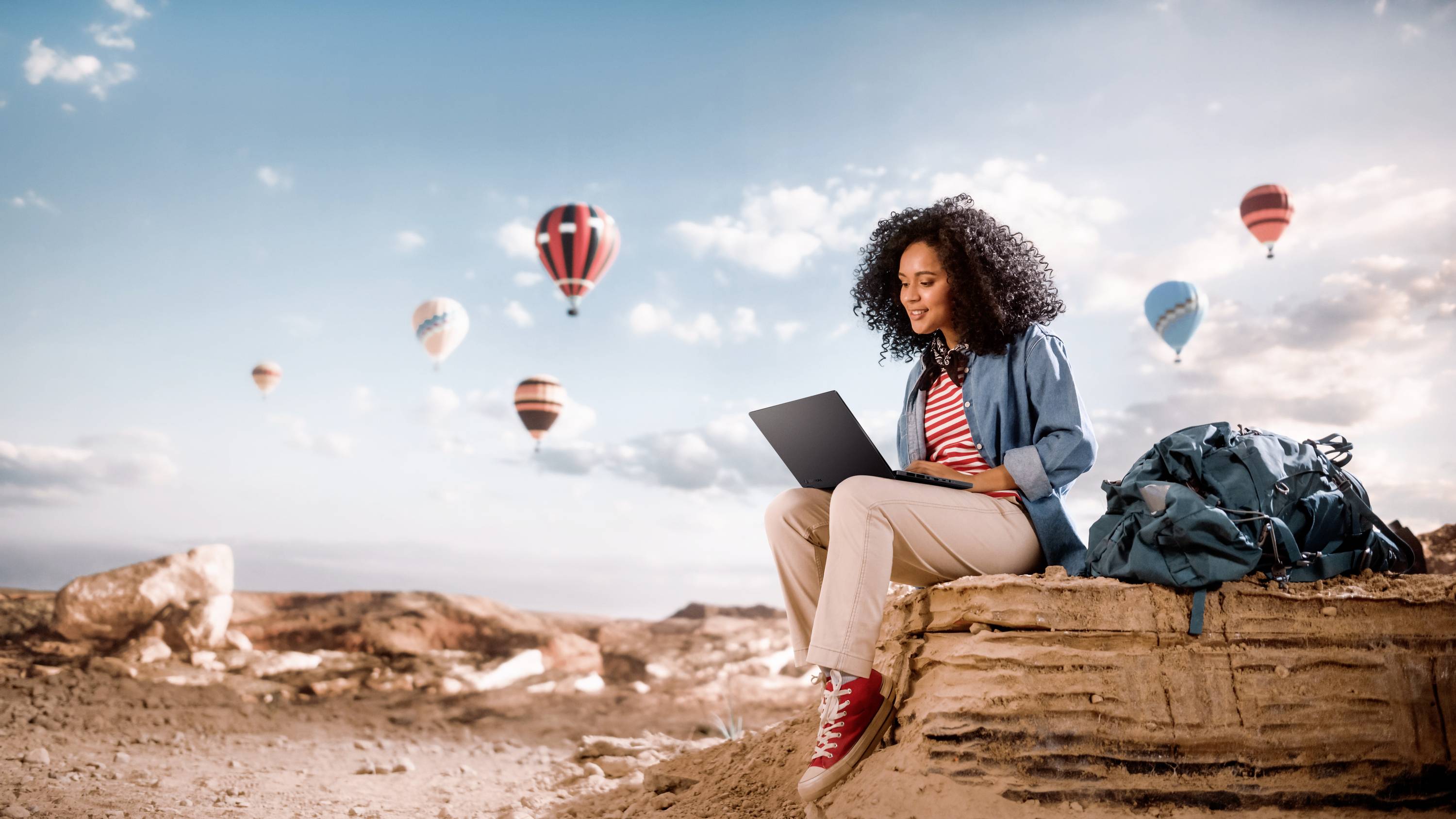 Eine Frau sitzt mit einem Laptop auf einem Felsen in einer Wüstenlandschaft, während bunte Heißluftballons am Himmel schweben.