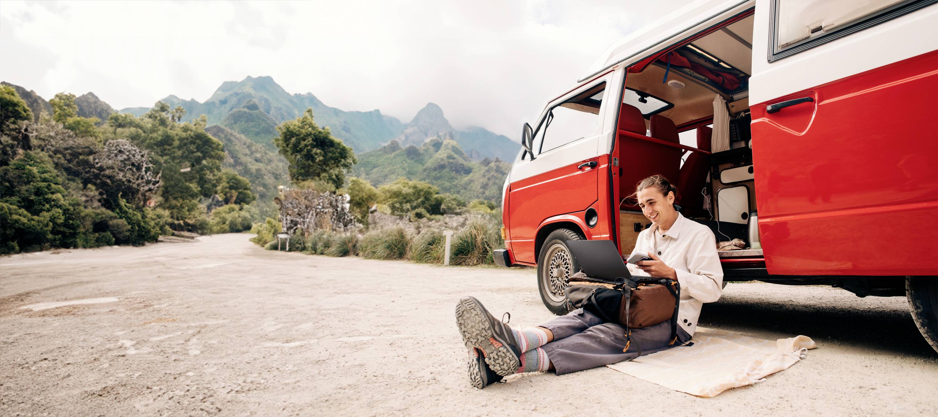 Ein Mann sitzt auf einer Decke neben einem roten Van vor einer Bergkulisse und benutzt ein Tablet. Er wirkt entspannt und zufrieden.