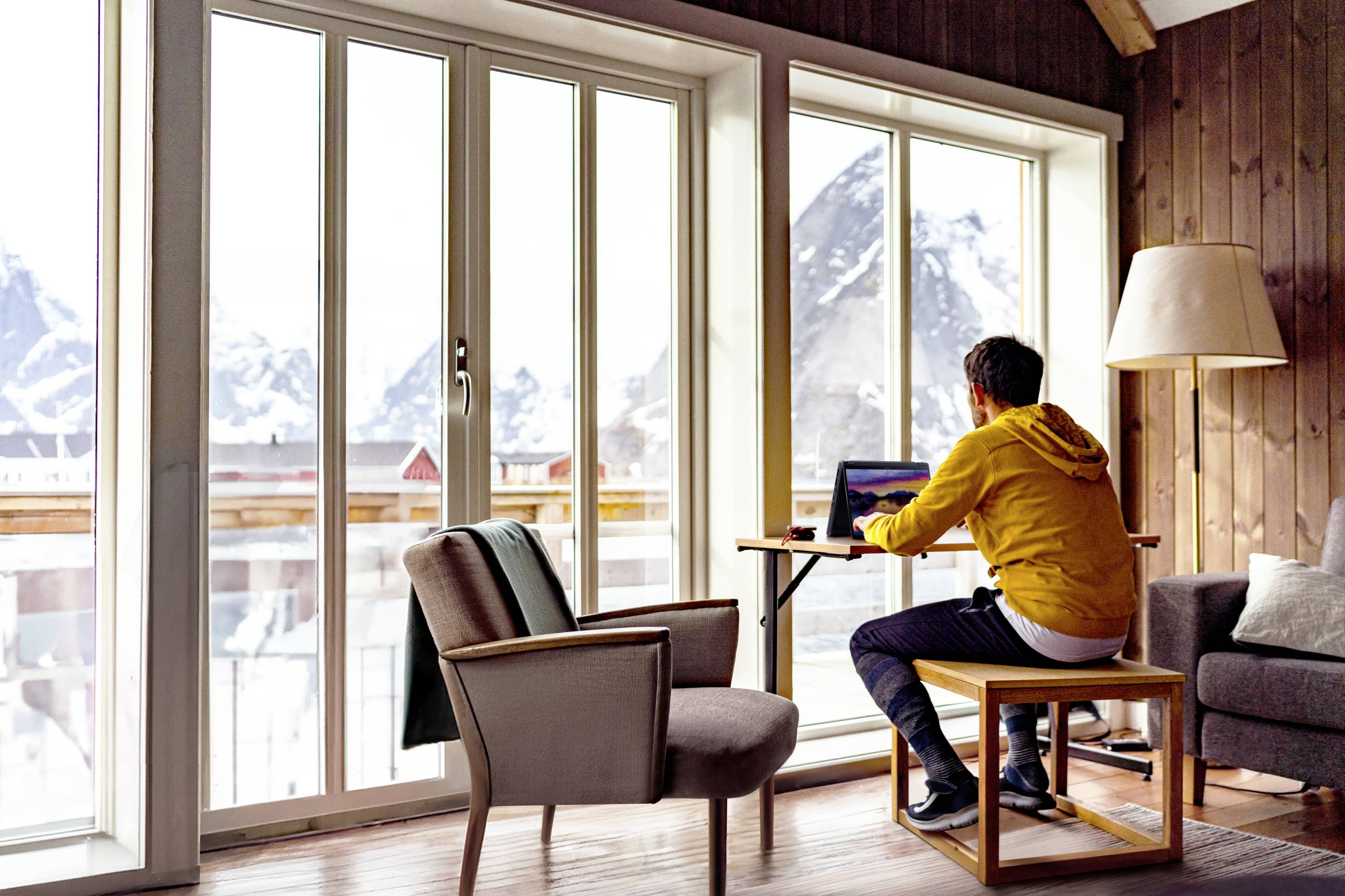 Person sitzt an einem Tisch mit Laptop in einem gemütlichen Raum mit großen Fenstern, durch die schneebedeckte Berge sichtbar sind.