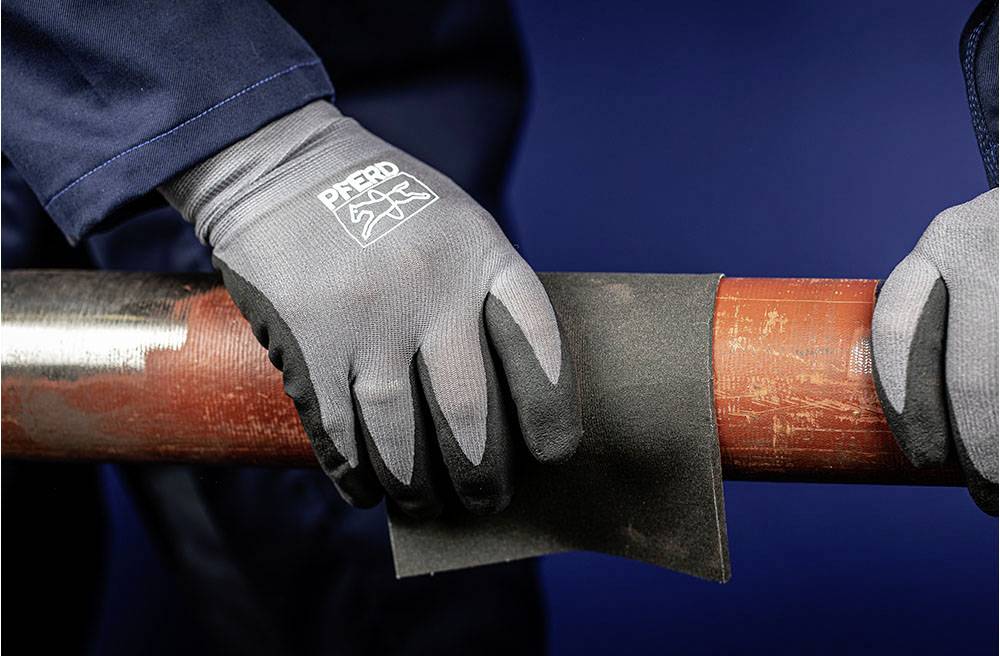 Zwei Hände mit grauen Arbeitshandschuhen schleifen ein rostiges Metallrohr mit Schleifpapier, Hintergrund in dunklem Blau.