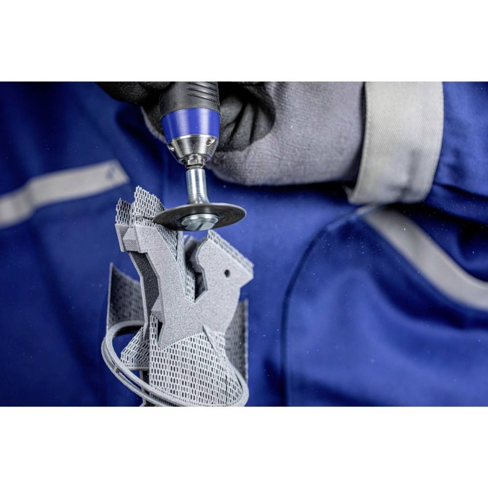 Eine Person schleift präzise eine graue Metallstruktur mit einem rotierenden Werkzeug, im Hintergrund blaues Arbeitsoutfit.