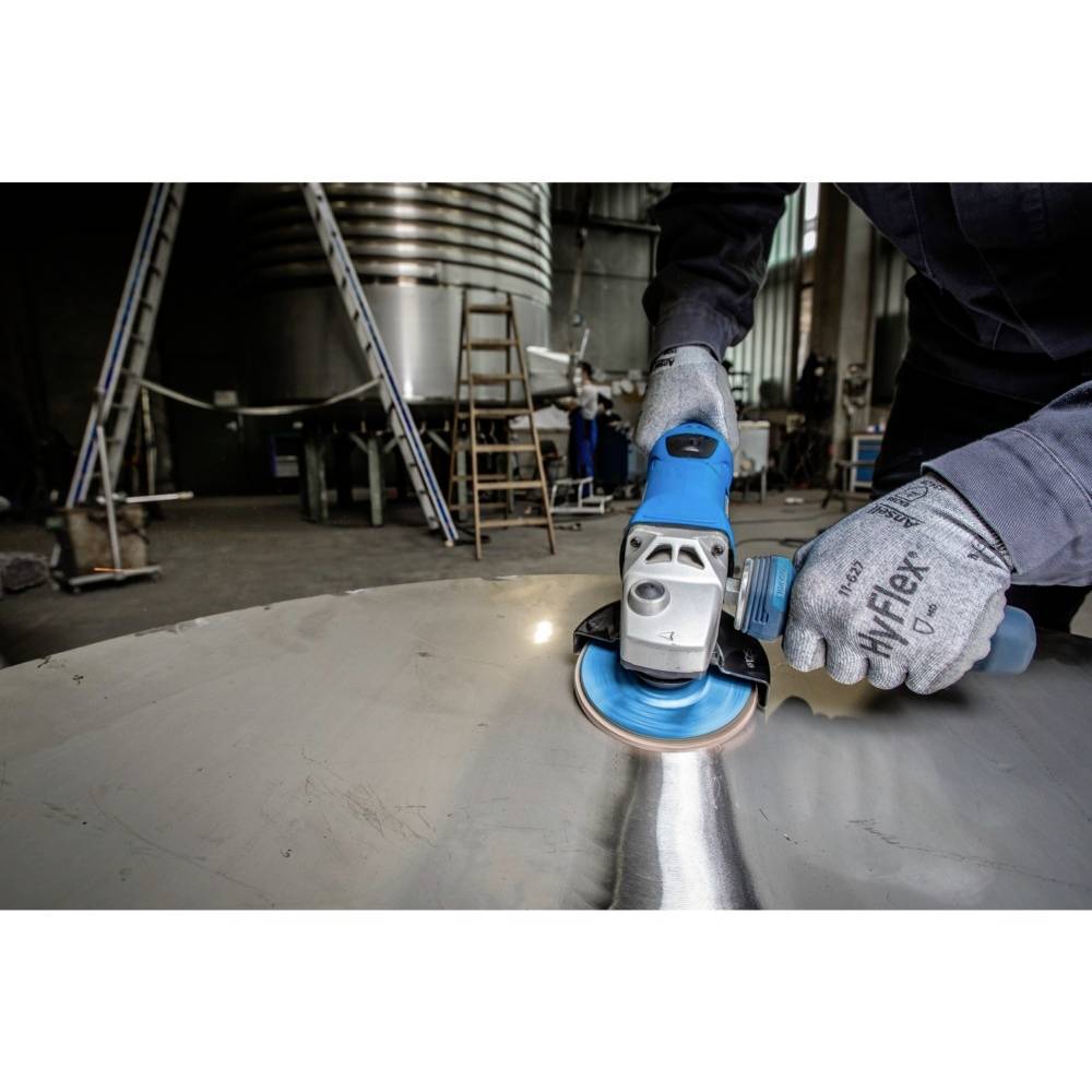 Eine Person schleift eine Metalloberfläche in einer Werkstatt mit einem Handwerkzeug. Im Hintergrund sind Leitern und Maschinen.