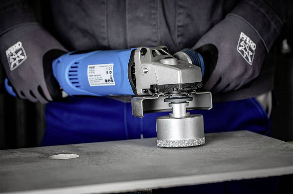 Eine Person schleift mit einem elektrischen Schleifgerät die Kante einer Metallplatte ab. Es werden Schutzhandschuhe getragen.