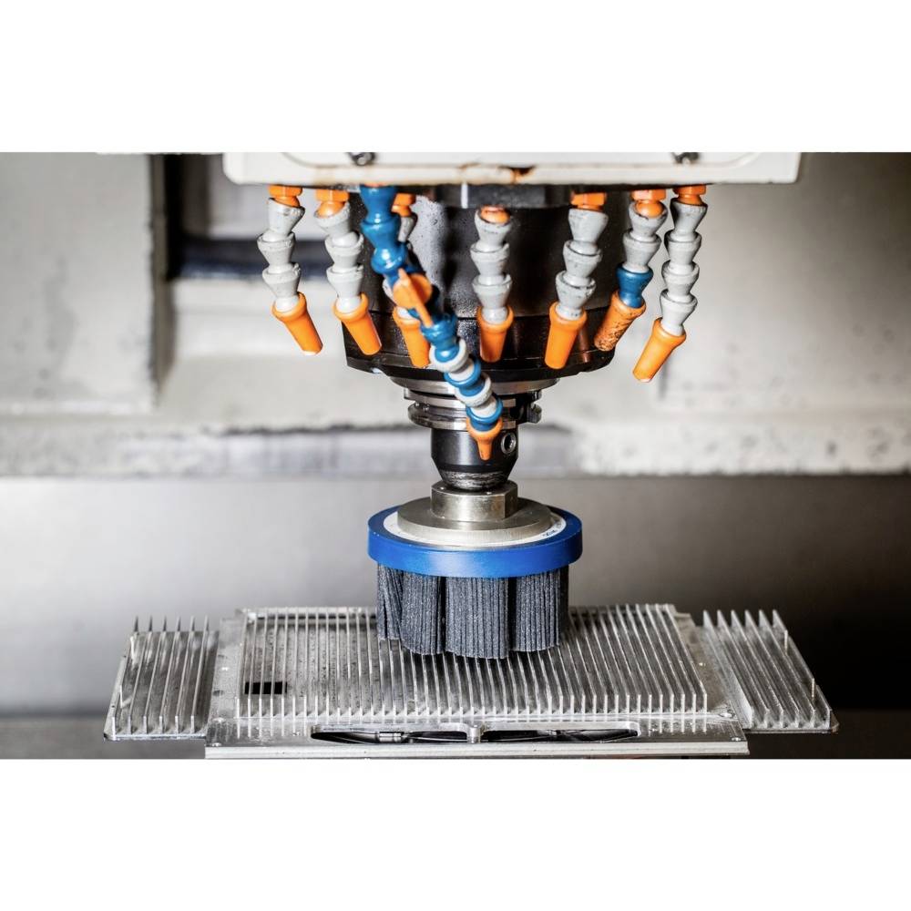 Industriemaschine mit rotierenden Bürsten und farbigen Düsen, die auf Metallteil arbeitet.