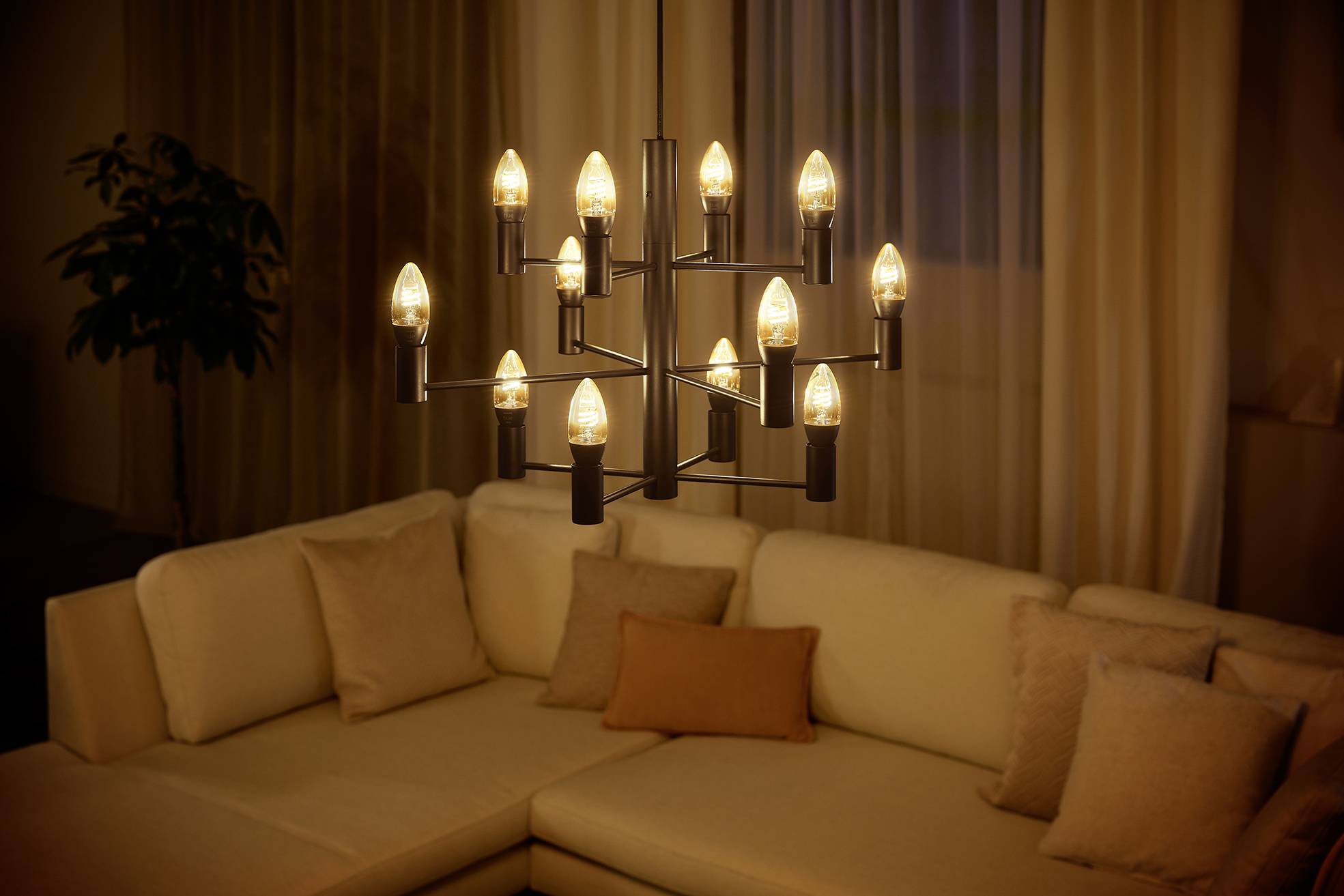 Ein Wohnzimmer mit einem cremefarbenen Ecksofa, auf dem mehrere Kissen liegen. Darüber hängt ein moderner Kronleuchter mit warmem Licht.