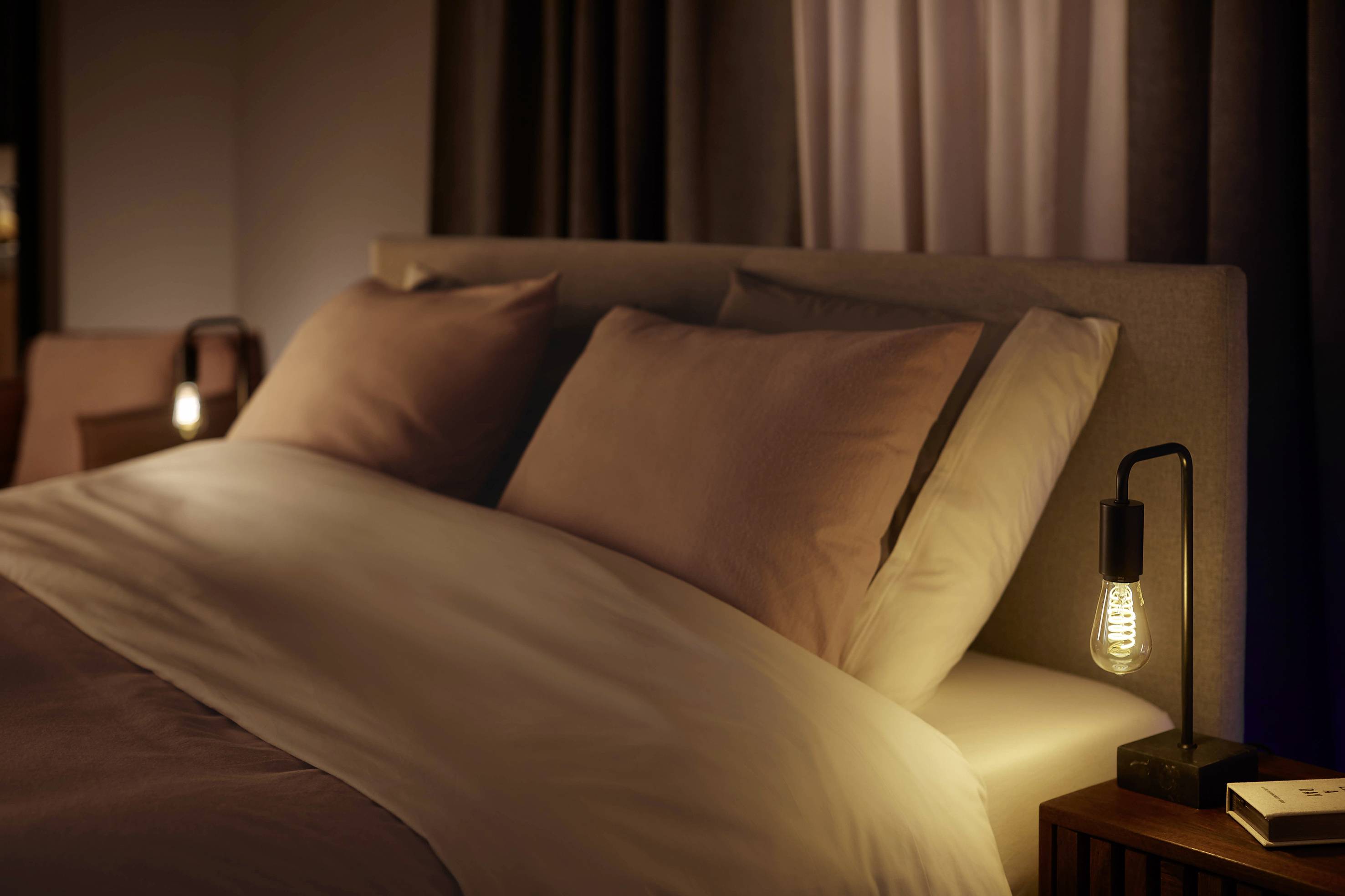 Bett mit weißen Laken und warm beleuchteten Nachttischlampen in einem gemütlichen, abgedunkelten Raum.