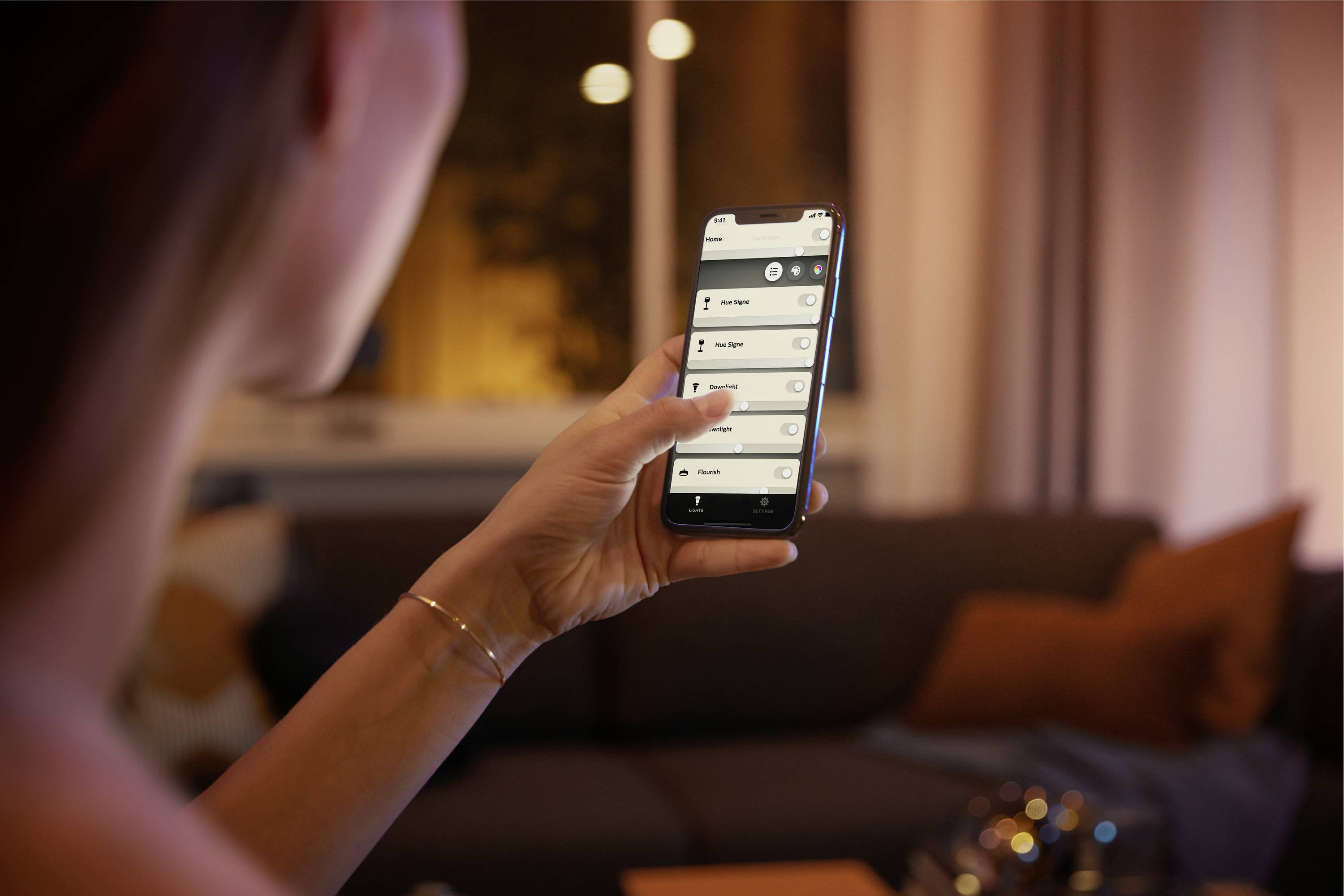 Eine Person bedient ein Smartphone, um smarte Lichter im Wohnzimmer zu steuern. Im Hintergrund sind ein Sofa und Fenster erkennbar.