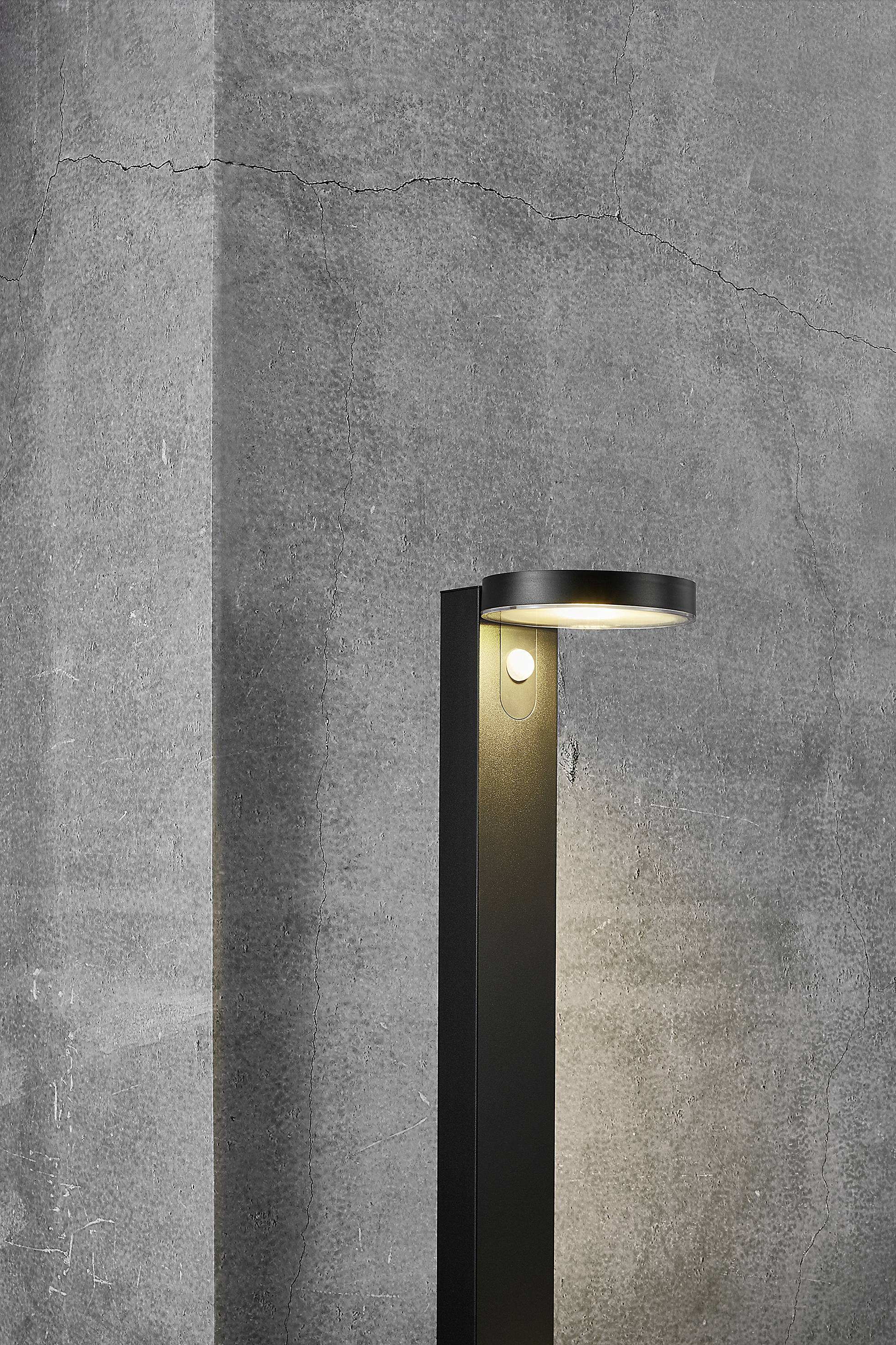 Eine moderne schwarze Stehlampe leuchtet gegen eine graue Betonwand. Die Lampe hat ein schlichtes, minimalistisches Design.