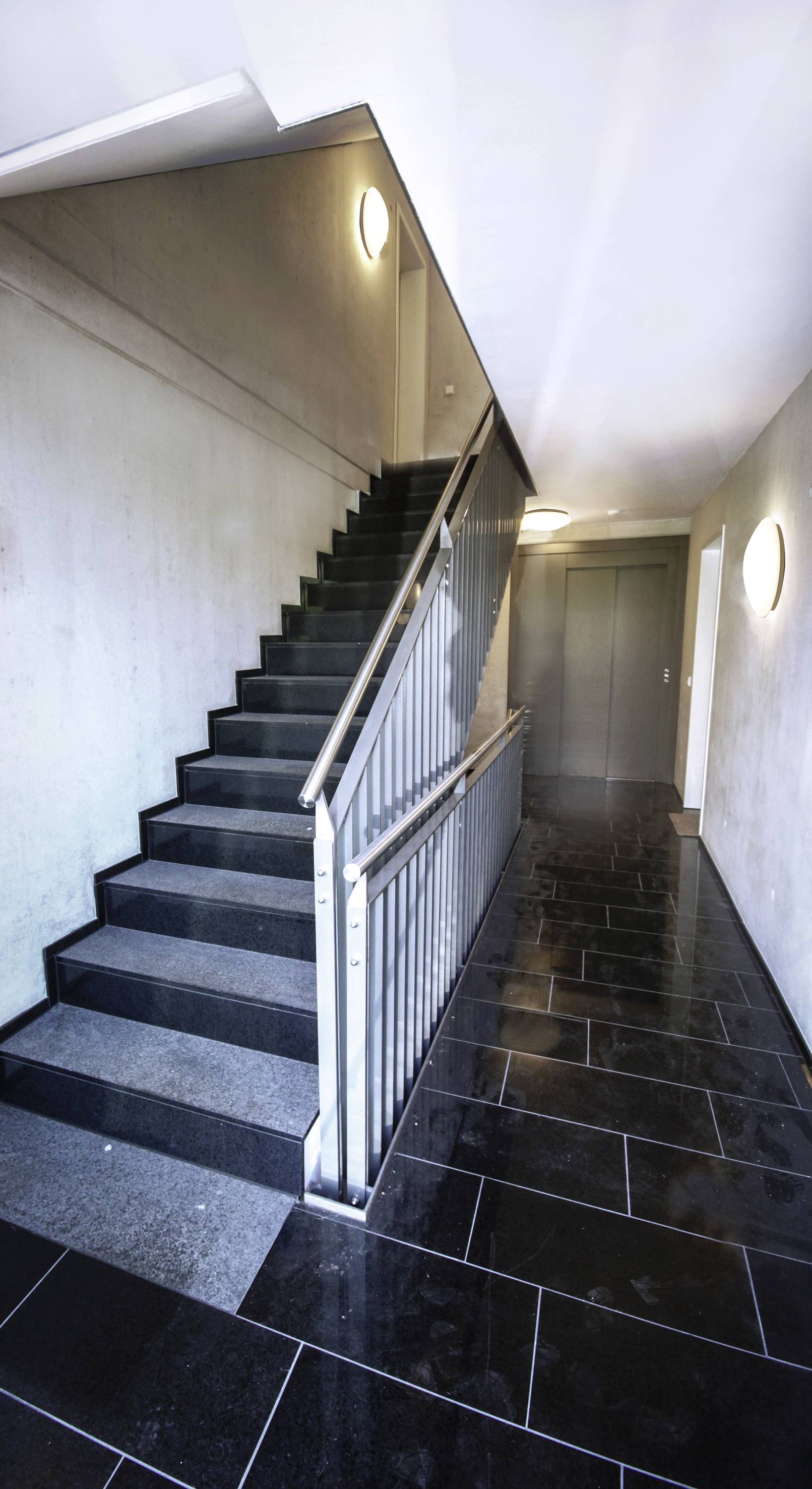 'Treppenhaus mit dunklen Fliesen und grauen Wänden, links eine Treppe mit Metallgeländer, geradeaus eine geschlossene Tür.'