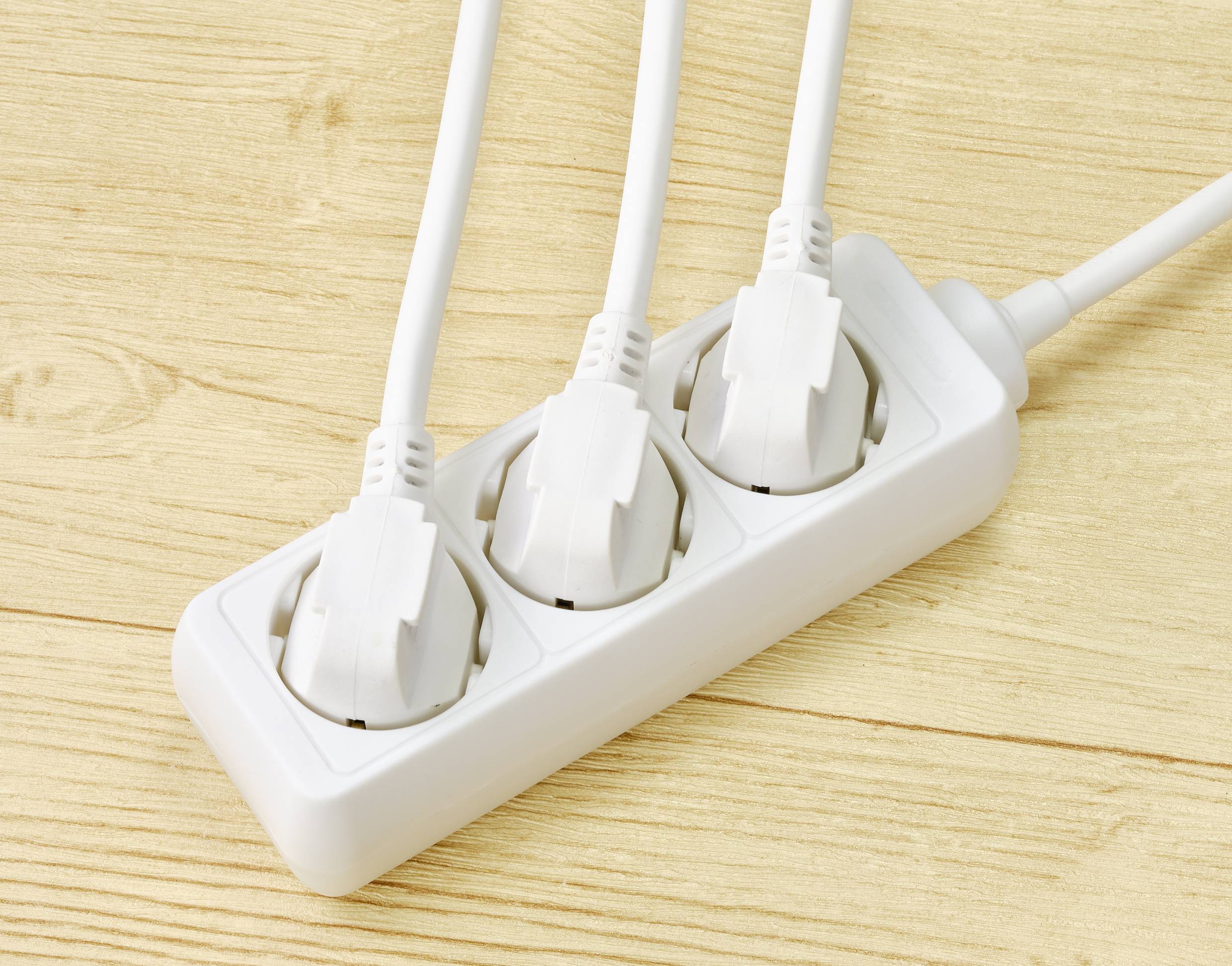 Ein weißer Mehrfachstecker mit drei eingesteckten weißen Steckern auf einem Holzuntergrund.