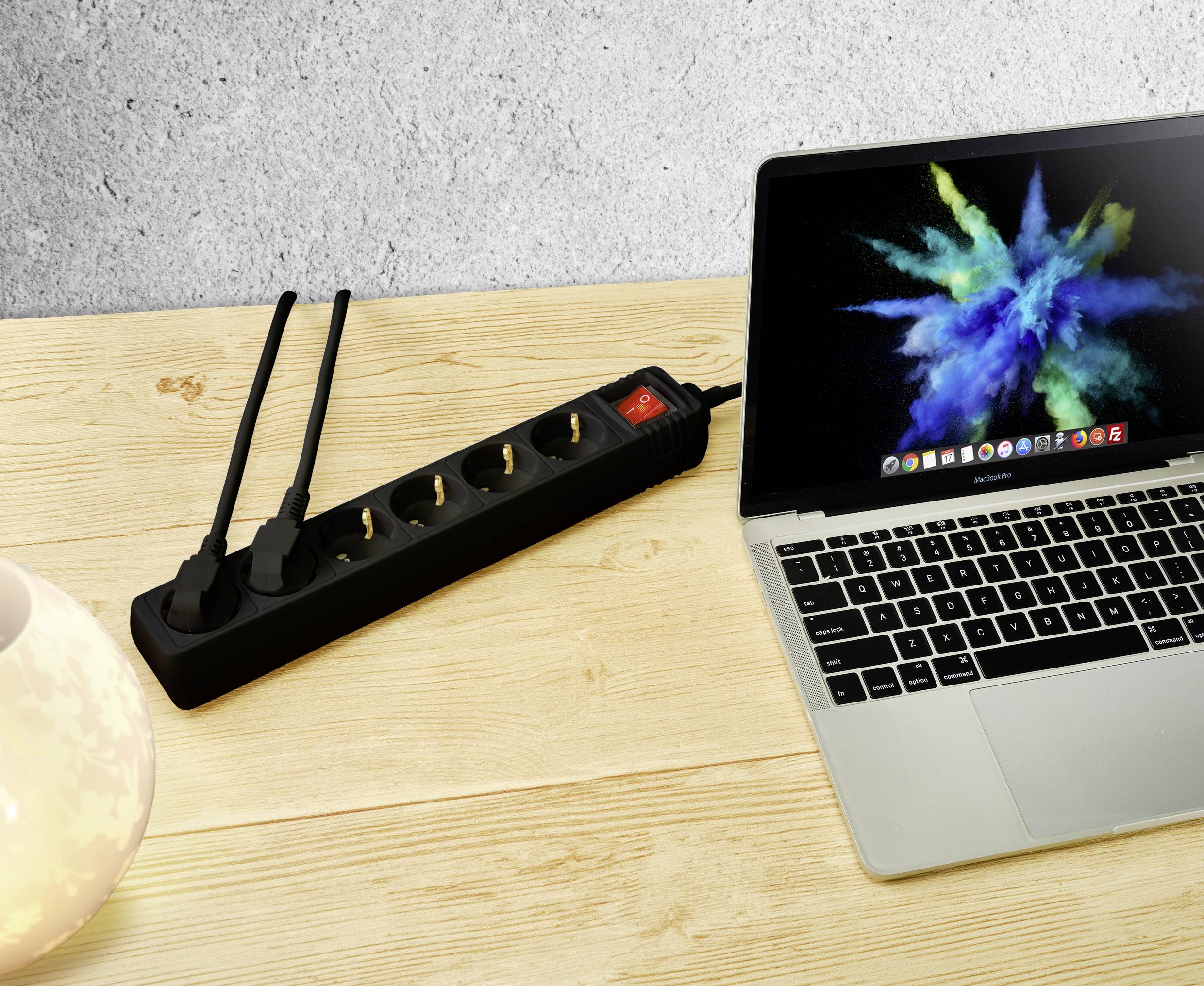 Laptop auf Holztisch neben einer Steckdosenleiste mit zwei eingesteckten Netzsteckern. Laptop zeigt bunte Bildschirmgrafik.