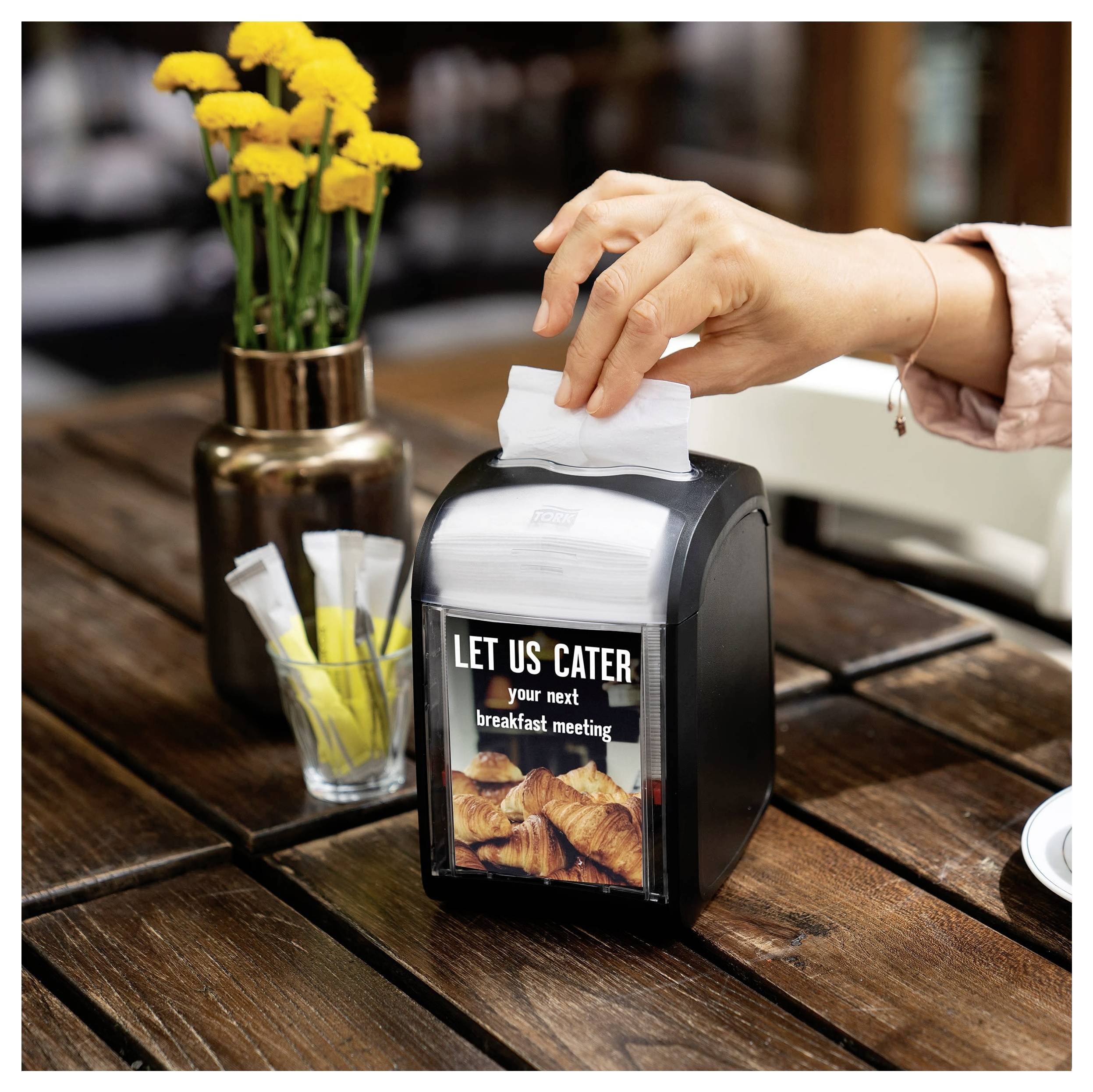 Eine Hand legt eine Serviette in einen Tischausgeber, auf dem „LET US CATER your next breakfast meeting" steht, neben einer Vase mit gelben Blumen.