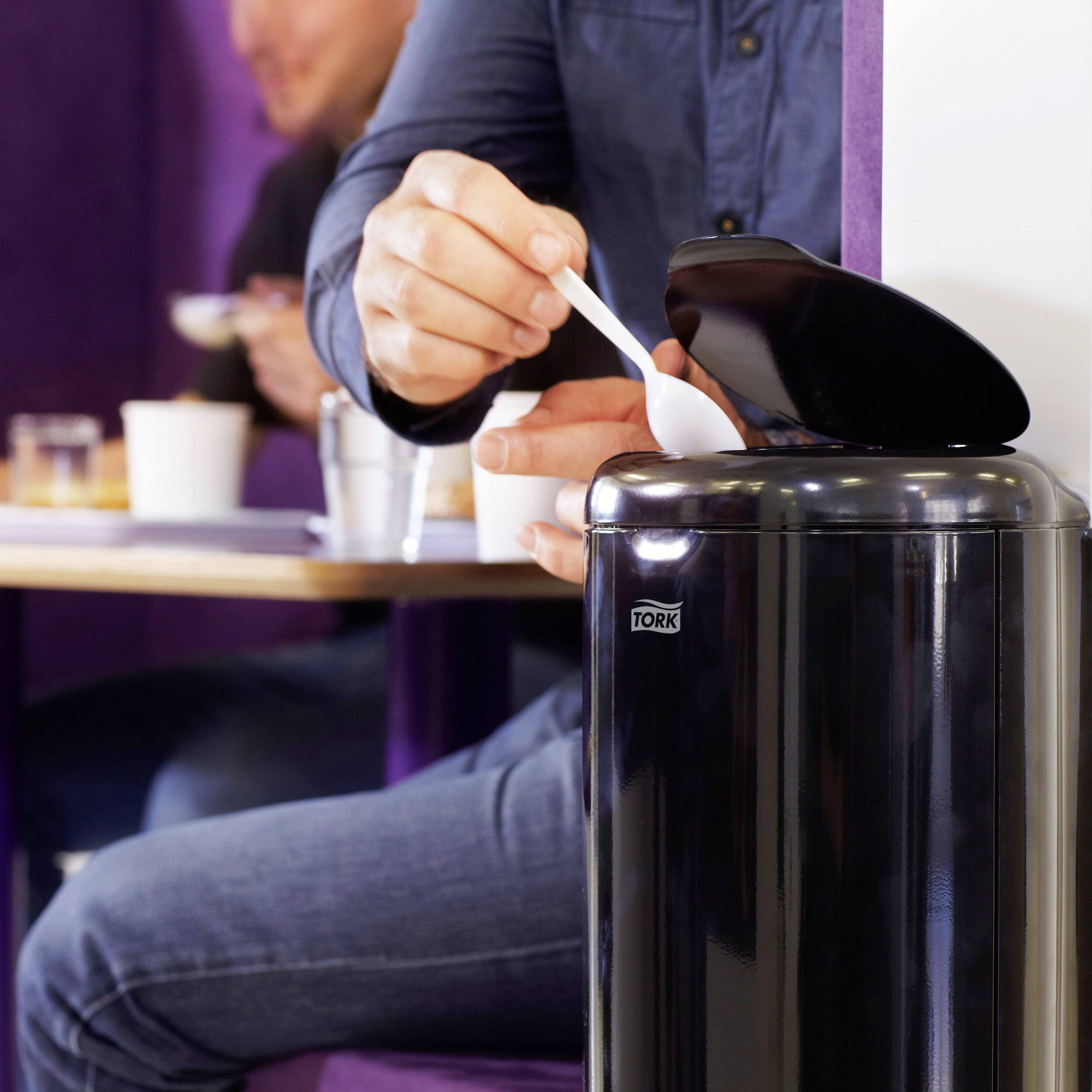 Eine Person entsorgt einen weißen Löffel in einen schwarzen Abfalleimer in einem Café. Weitere Personen sitzen im Hintergrund.