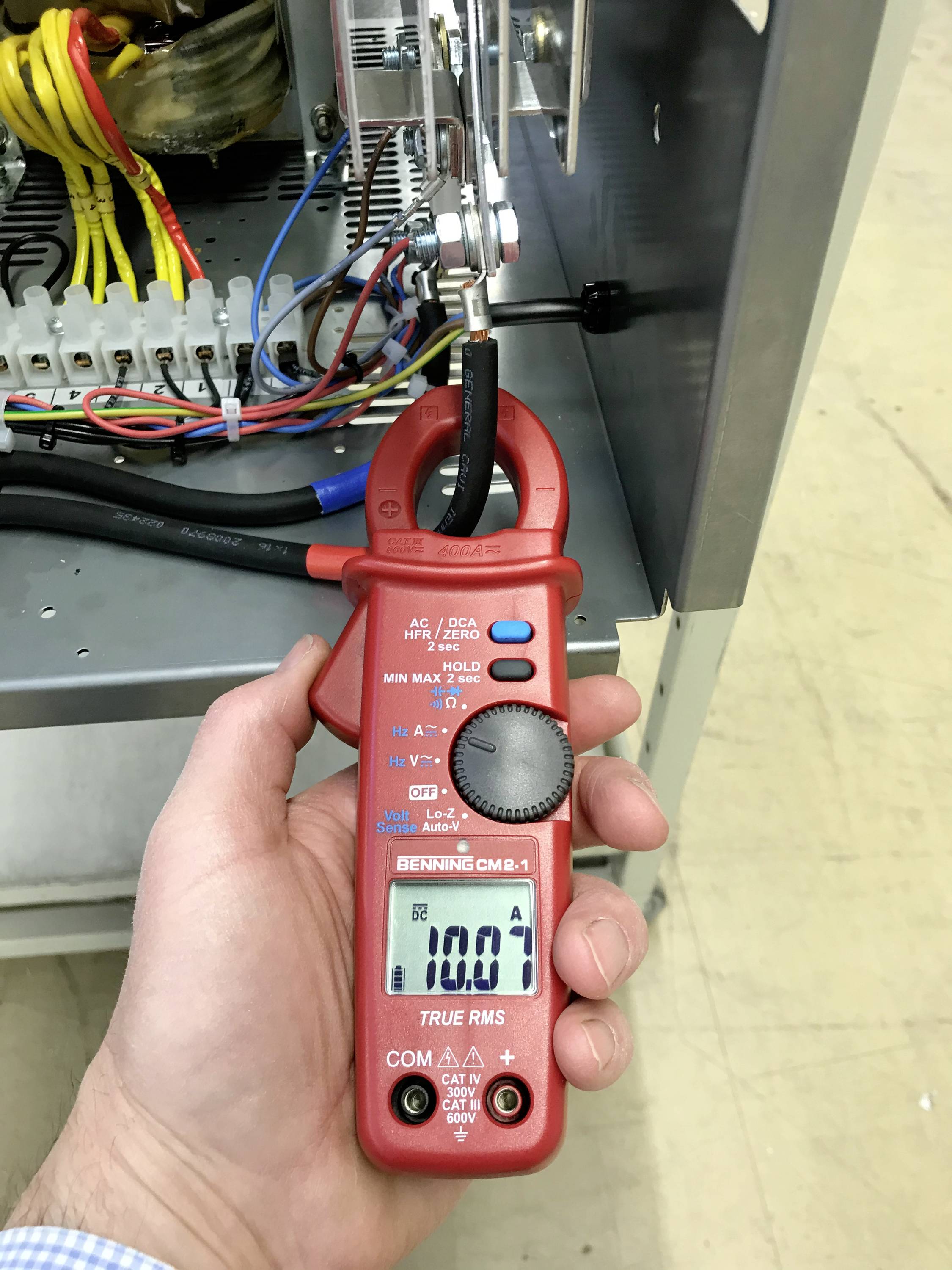 Eine Hand hält ein rotes Strommessgerät. Es zeigt '100,0 A' auf einem Display an und misst den Strom eines Kabels in einem Schaltschrank.