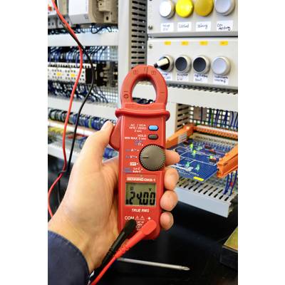 Person hält einen roten digitalen Spannungsmesser vor einem Schaltkasten. Das Display zeigt '24.0' an. Hintergrund mit Kabeln und Schaltungen.