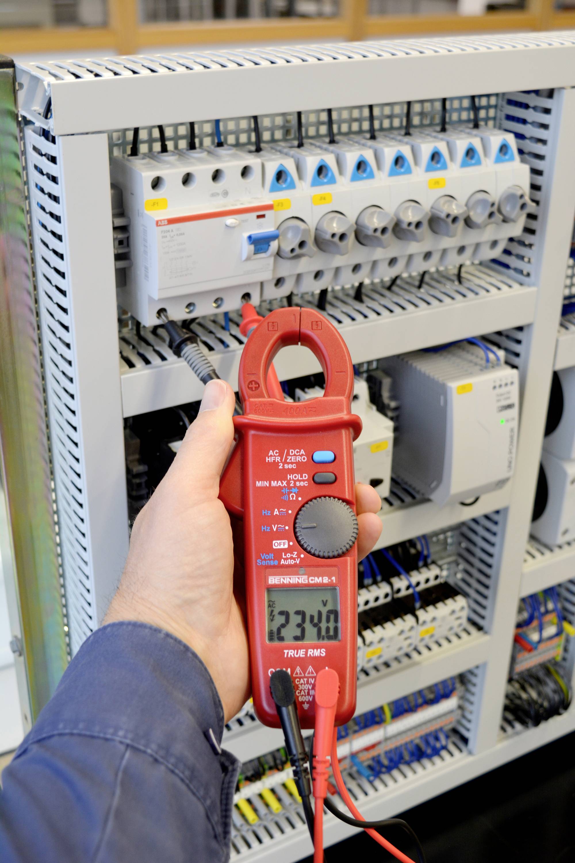 Eine Person misst mit einem roten Multimeter die Spannung in einem Schaltschrank. Das Display zeigt '234.0' Volt an.