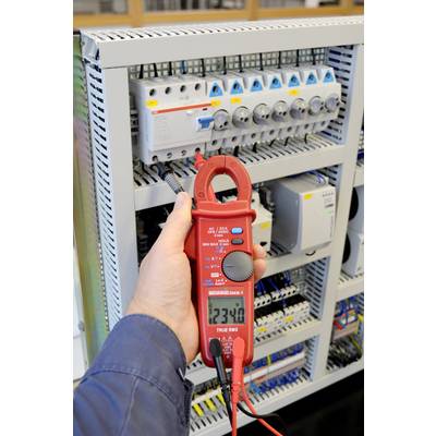 Eine Person misst mit einem roten Multimeter die Spannung in einem Schaltschrank. Das Display zeigt '234.0' Volt an.