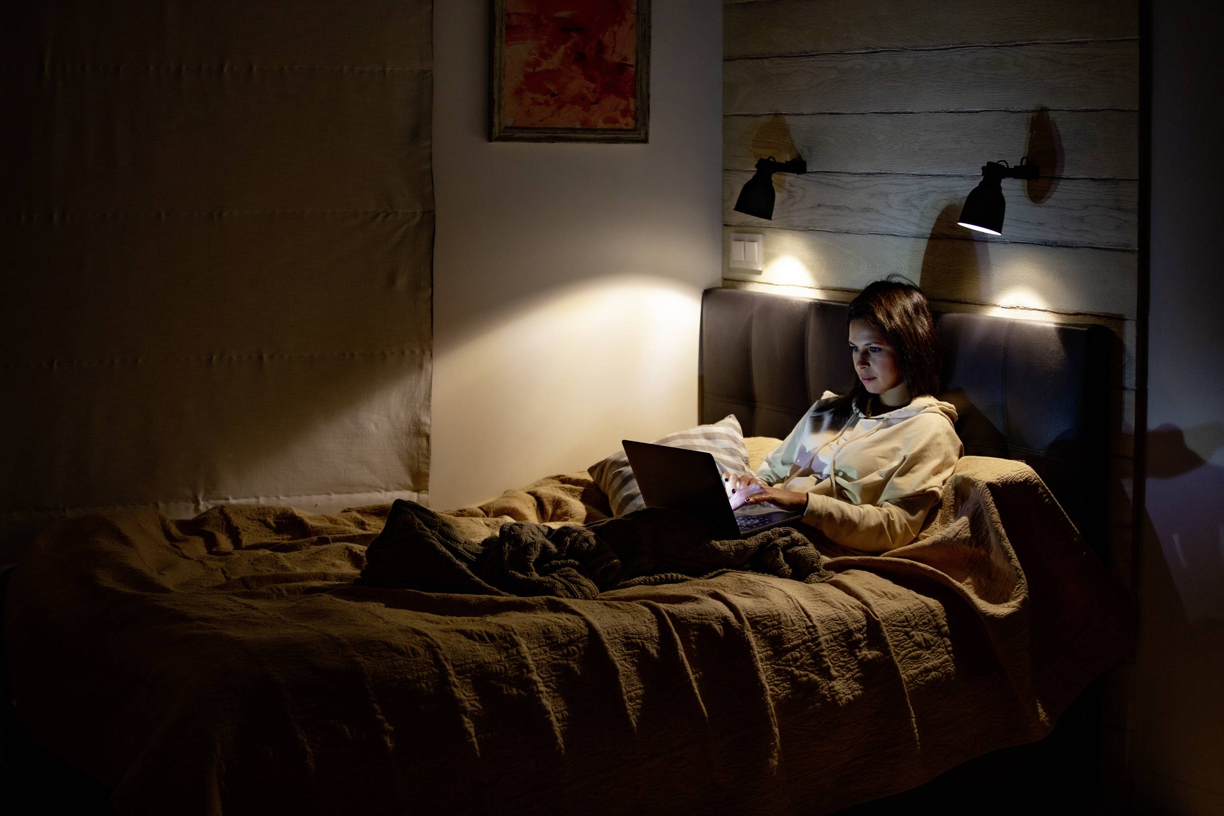 Frau sitzt in dunklem Raum auf dem Bett und arbeitet an einem Laptop, nur vom Bildschirmlicht und einer Nachttischlampe beleuchtet.