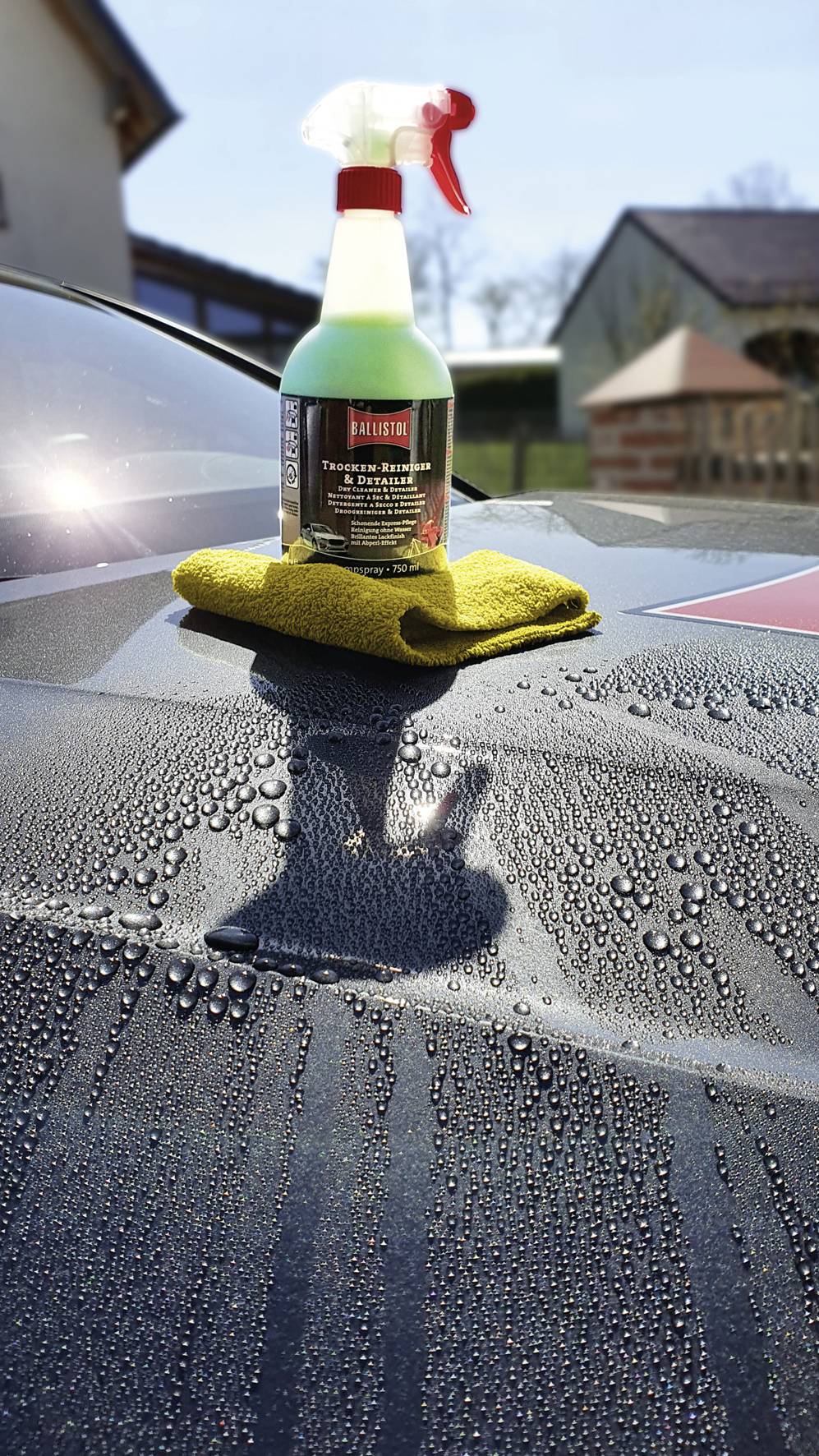 Eine Sprühflasche mit Autoreiniger steht auf einem gelben Mikrofasertuch, das auf der nassen Motorhaube eines Autos liegt.