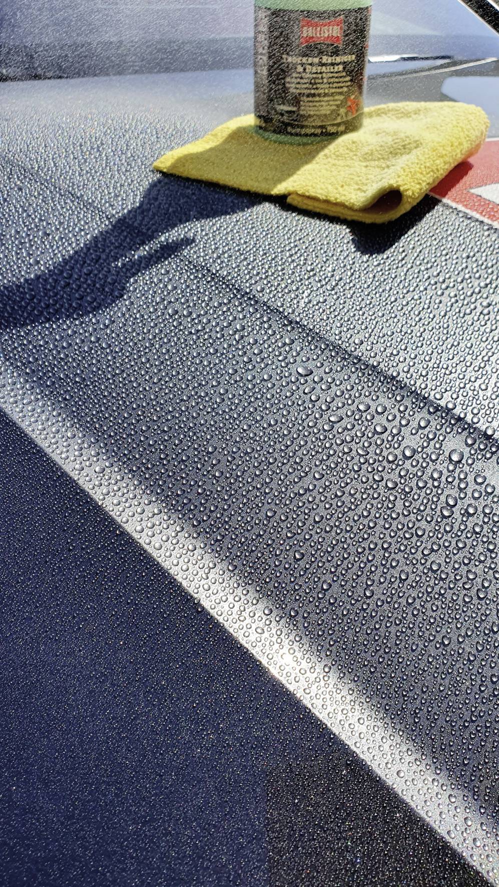 Ein Wachsbehälter und ein gelbes Tuch liegen auf einer mit Wassertropfen bedeckten Autooberfläche, bereit zur Autopflege.