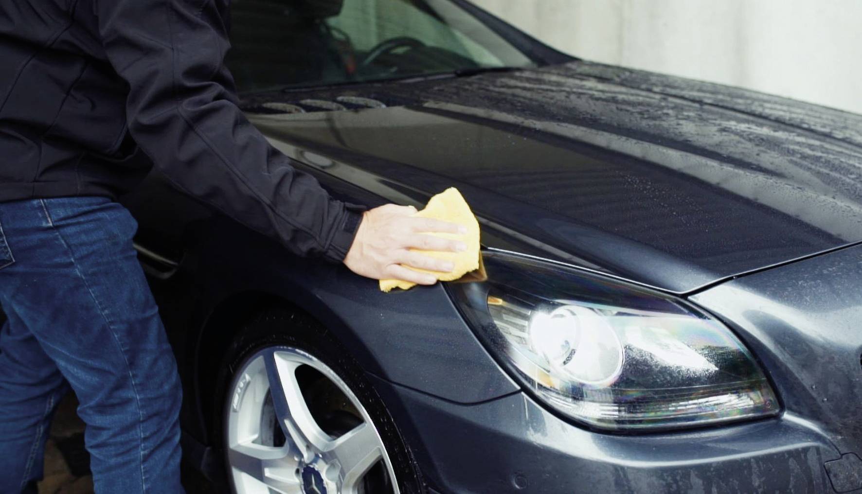 Person reinigt die Motorhaube eines Autos mit einem gelben Tuch. Das Auto ist dunkel und steht in einer Garage.