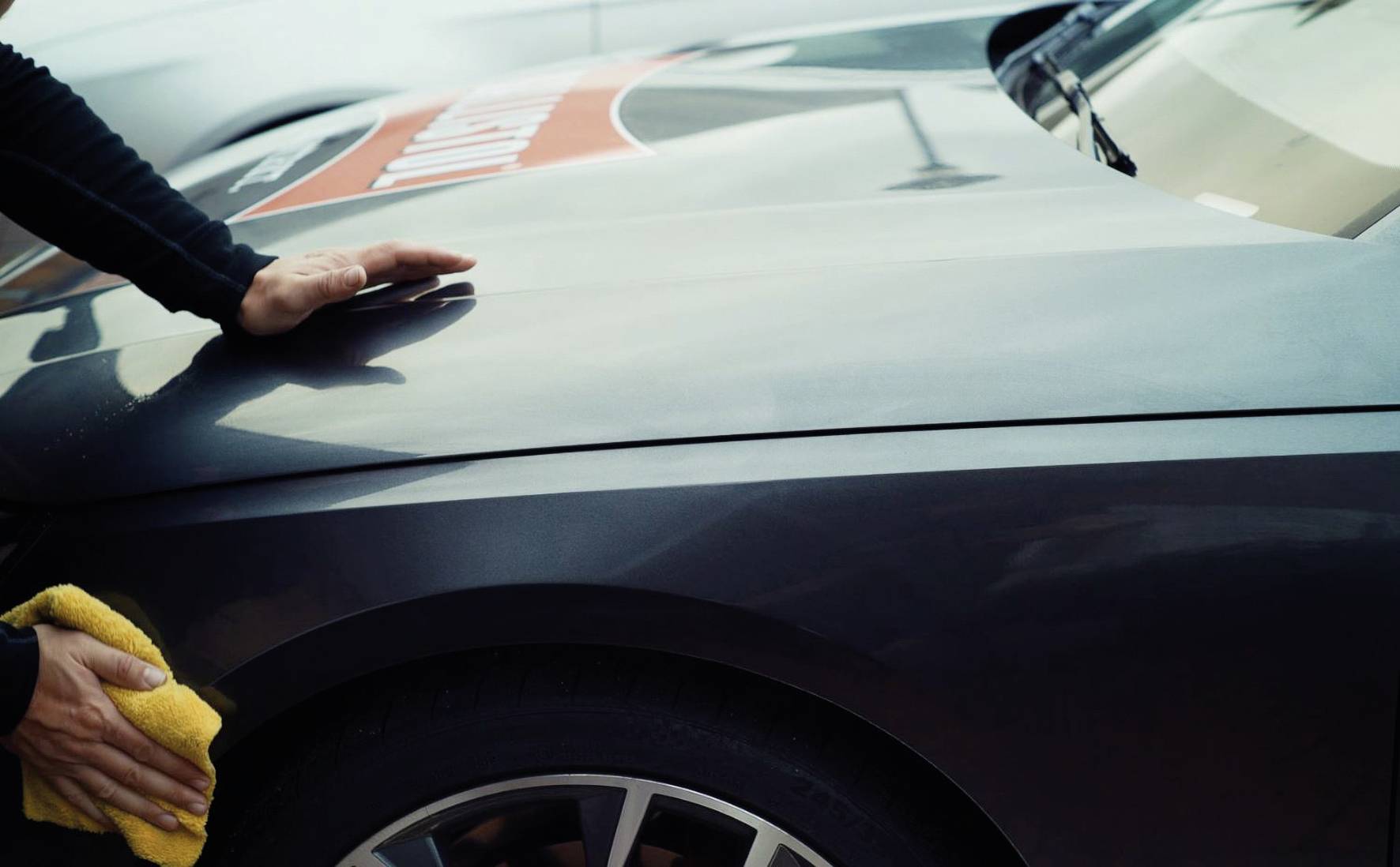 Eine Person poliert mit einem gelben Tuch den Kotflügel eines dunklen Autos. Hände und Tuch sind sichtbar, das Auto steht im Fokus.