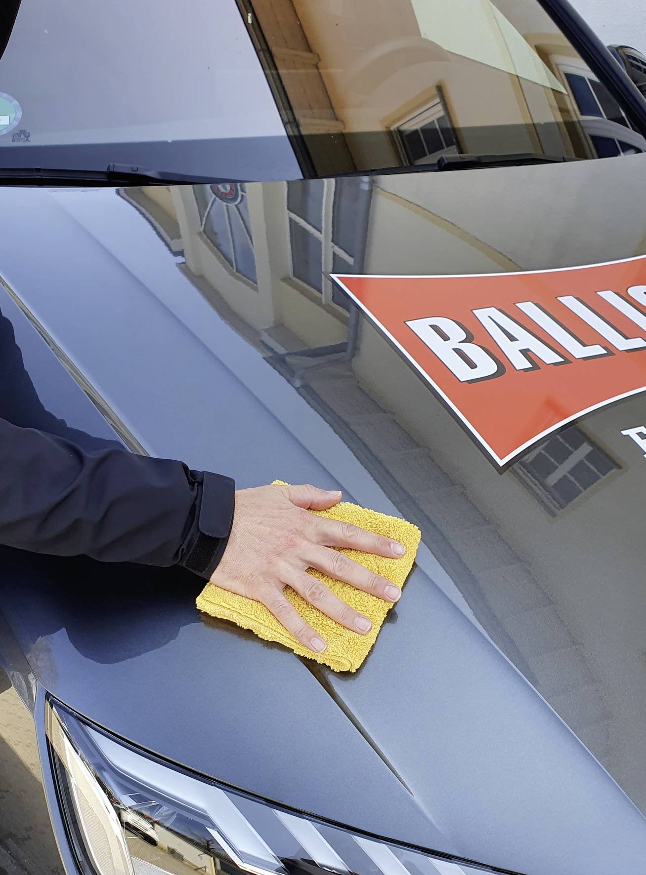 Person wischt mit einem gelben Tuch die Motorhaube eines Autos mit einem Aufkleber.