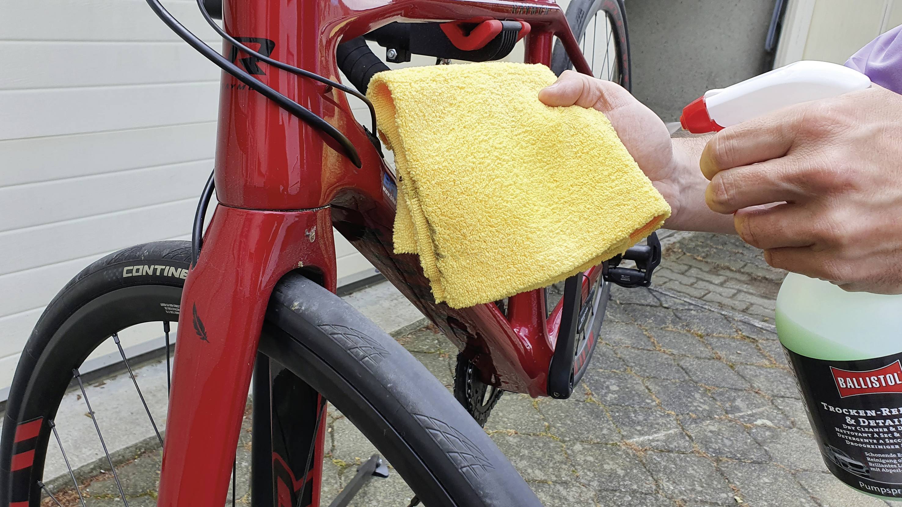 Person reinigt ein rotes Fahrrad mit einem gelben Tuch und einem Sprayreiniger. Das Fahrrad steht draußen auf einem gepflasterten Weg.