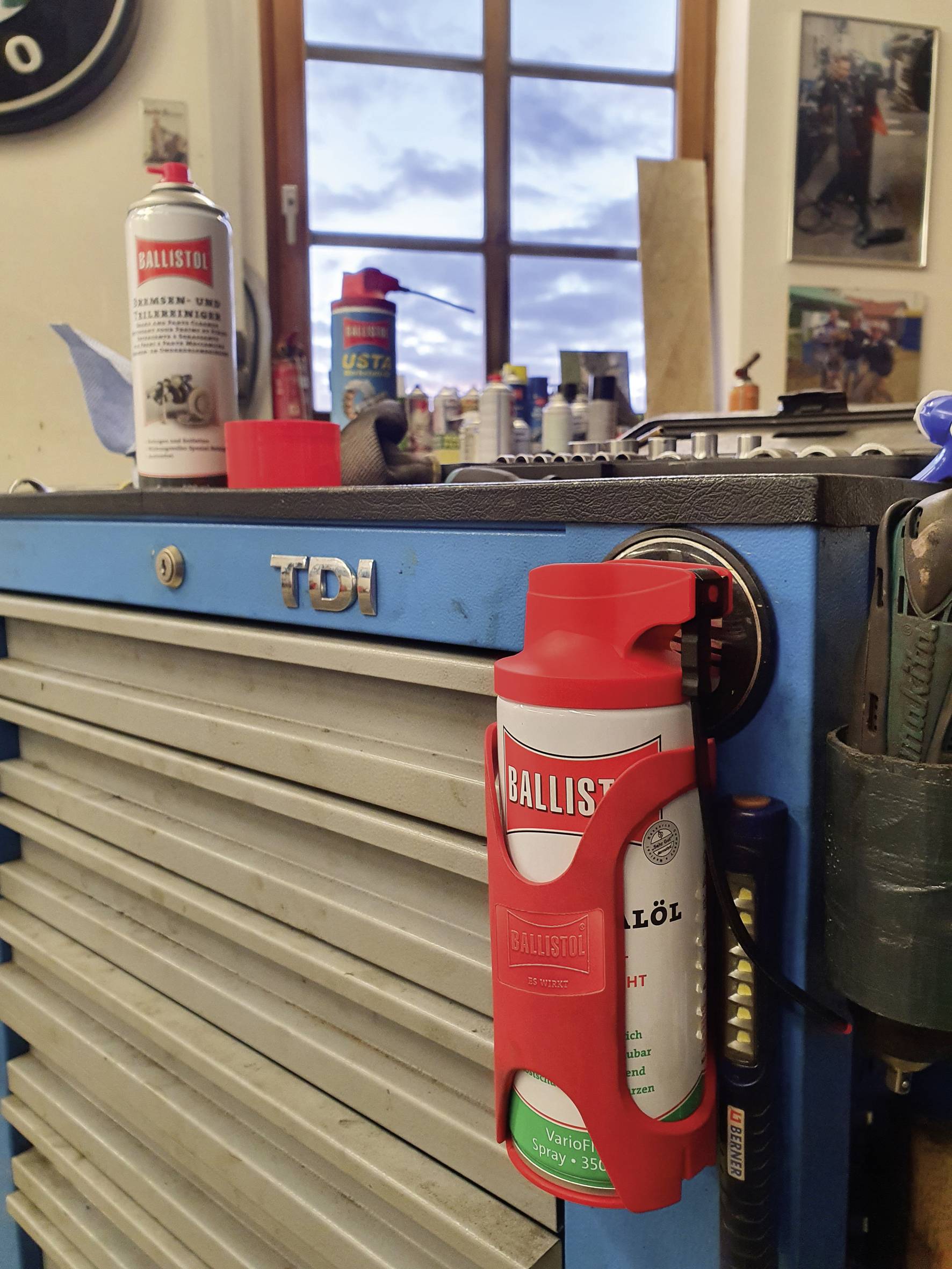 Ein Werkstattwagen mit einem TDI-Logo. Darauf stehen Spraydosen, und daneben hängt eine rote Spraydose an einem Halter. Im Hintergrund ein Fenster.