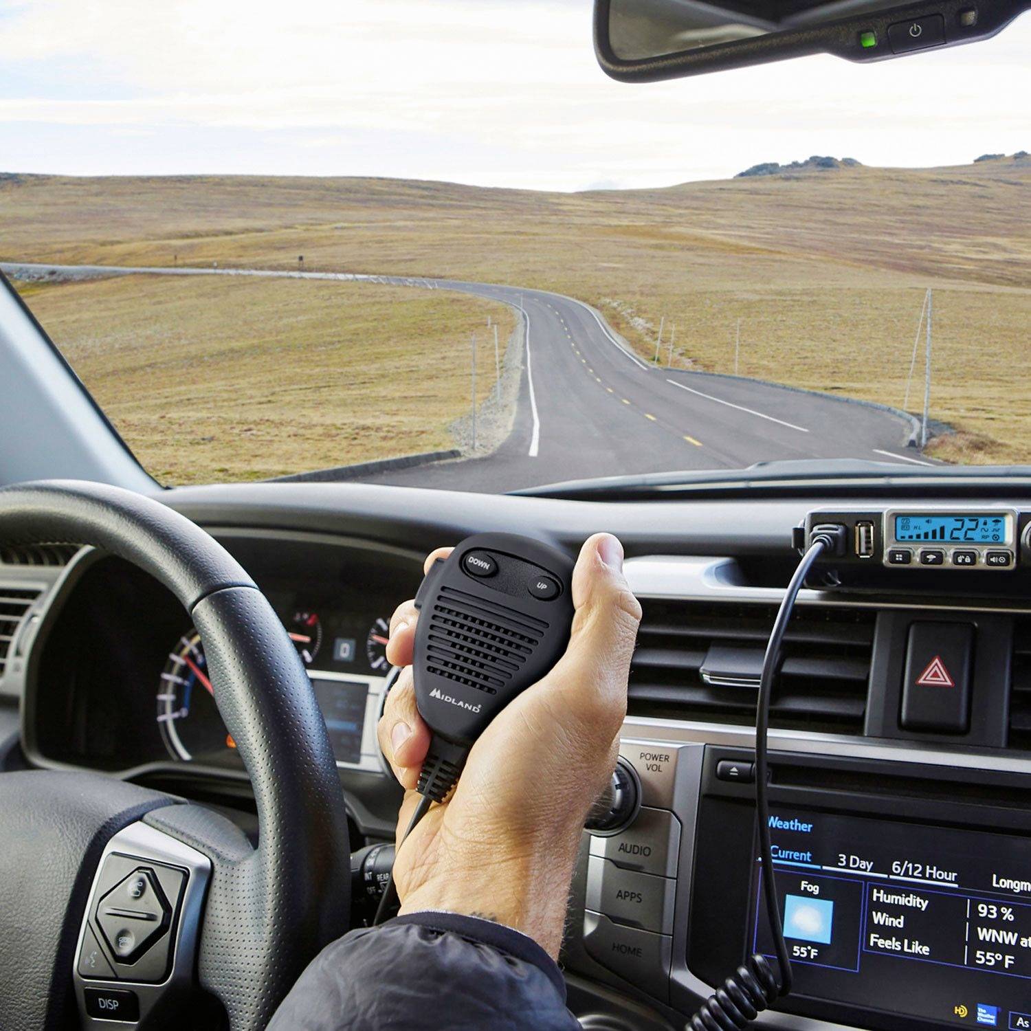 Blick aus einem Auto auf eine kurvige Straße. Eine Person hält ein Funkgerät. Verkehrsfreie, ländliche Landschaft im Hintergrund.