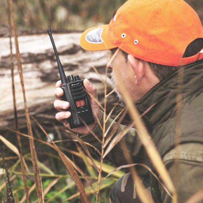 Ein Mann mit orangefarbener Kappe hält ein Funkgerät im Gras. Er scheint auf etwas in der Ferne zu achten, als ob er auf der Jagd sei.