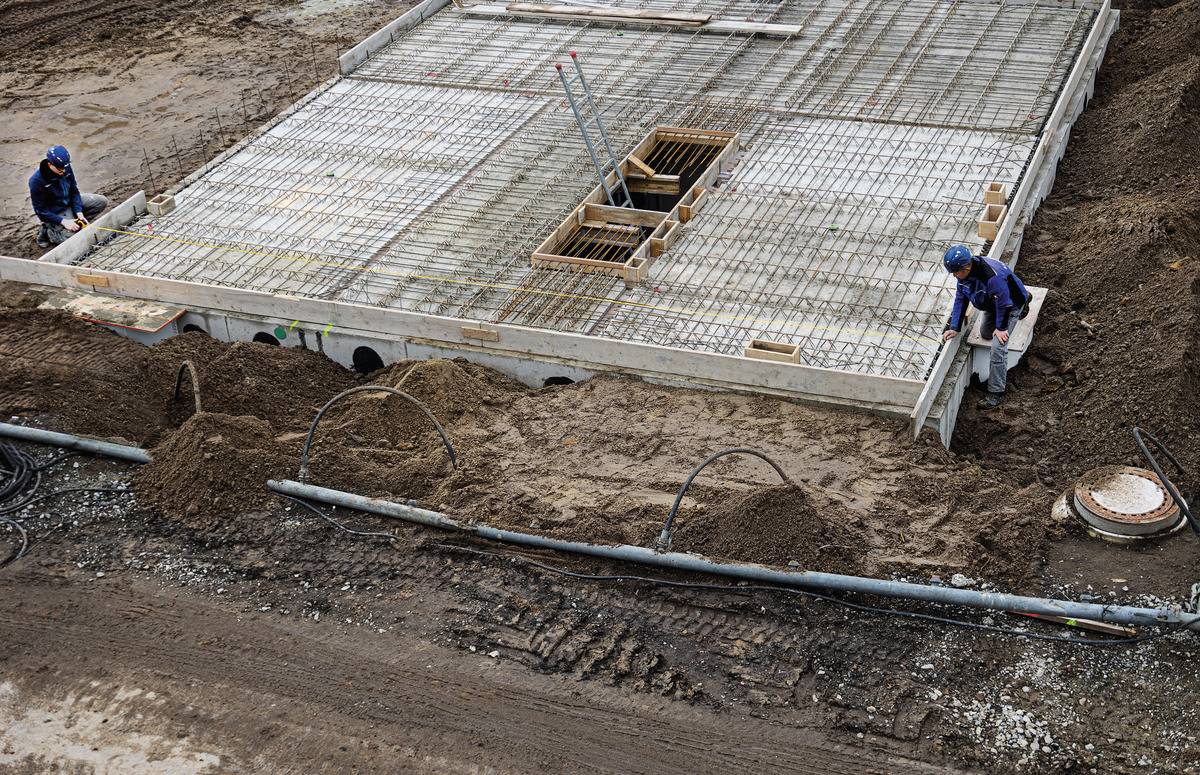 Eine Baustelle mit zwei Bauarbeitern, die an einem Fundament arbeiten. Der Boden ist aus Beton mit freiliegender Bewehrung.