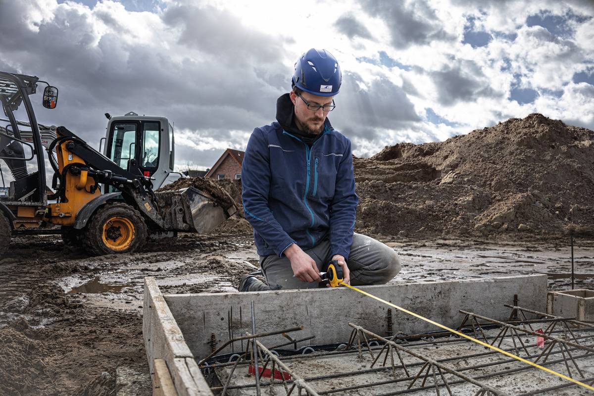 'Ein Bauarbeiter mit Helm misst mit einem Maßband ein Betonfundament auf einer Baustelle. Im Hintergrund ein Bagger und Erdaufschüttungen.'