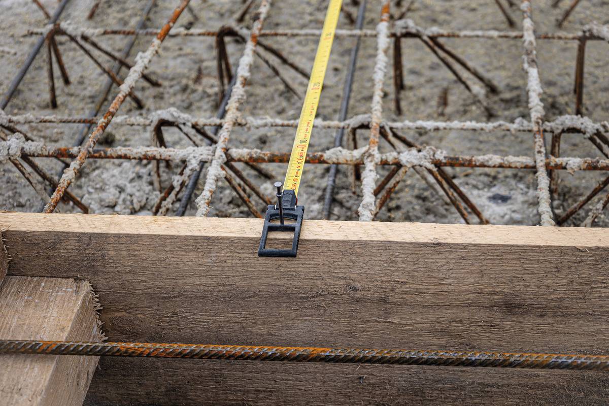 Ein Maßband liegt über einem Holzbrett vor einem Stahlgitter auf einer Baustelle, um die Größe für das Gießen von Beton zu messen.