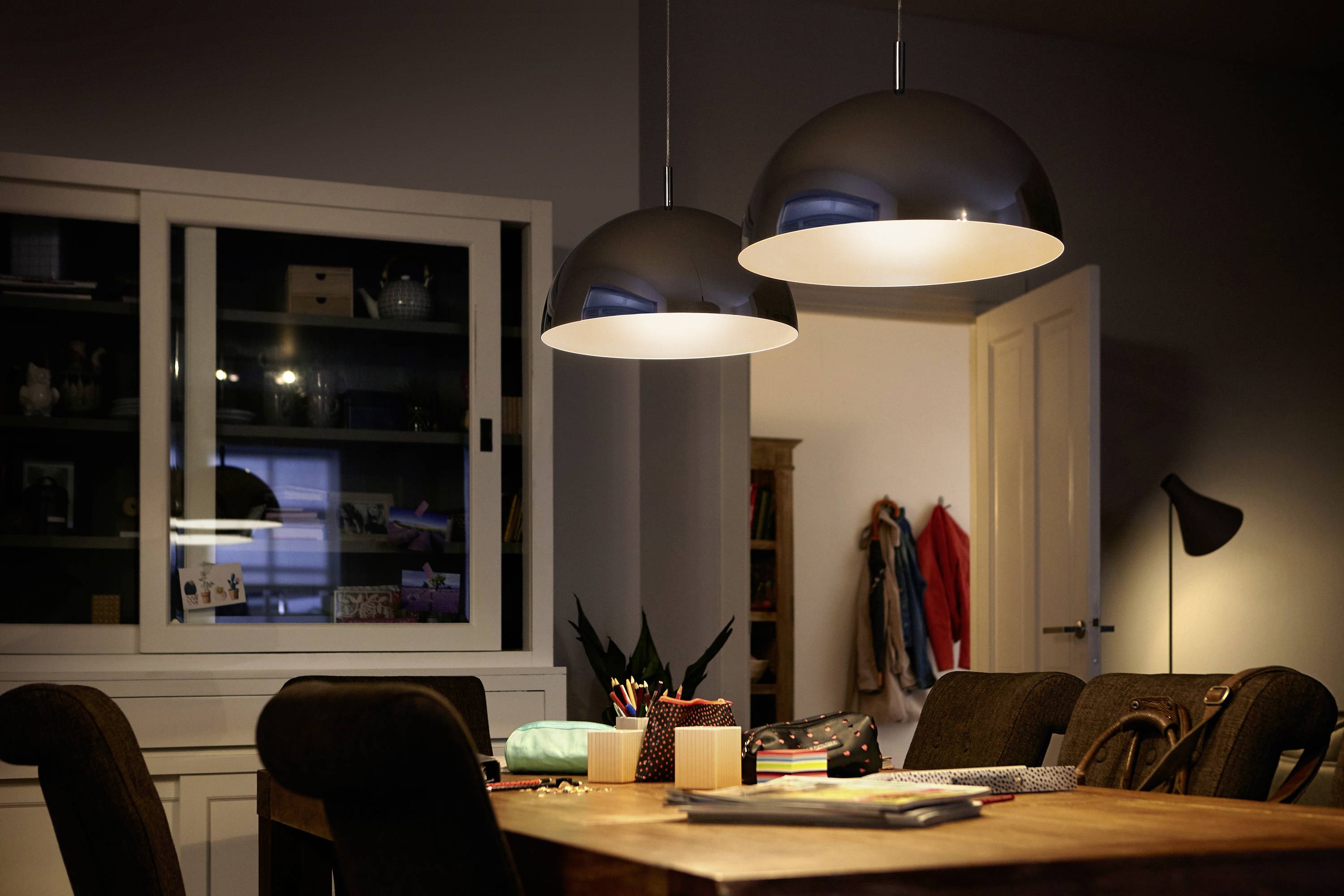 Ein Esszimmer mit einem Holztisch, umgeben von Stühlen. Zwei große, leuchtende Deckenlampen hängen über dem Tisch. Im Hintergrund ein Schrank und Garderobe.