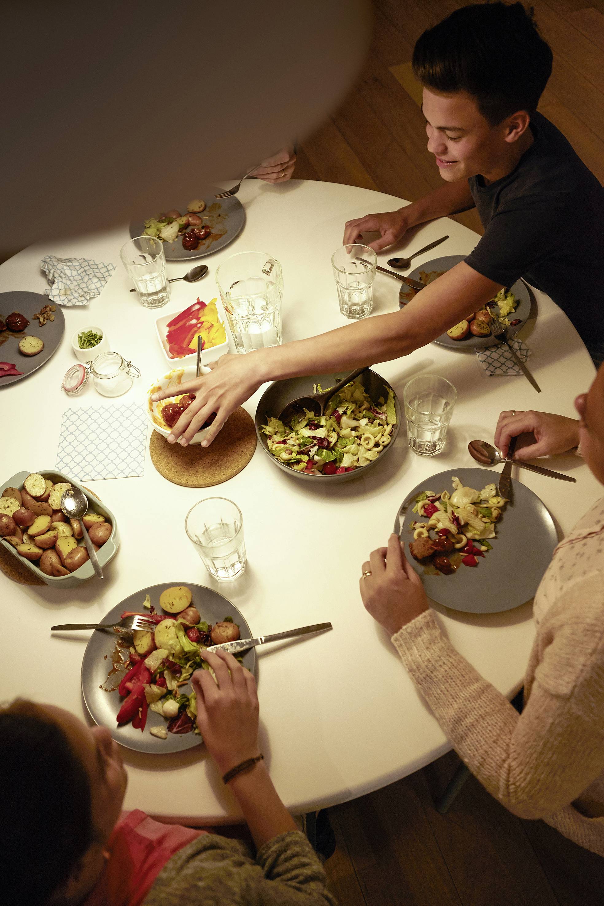 Personen sitzen um einen Tisch und essen gemeinsam. Auf dem Tisch stehen Teller mit Salat und gerösteten Kartoffeln. Eine Person reicht eine Schüssel weiter.