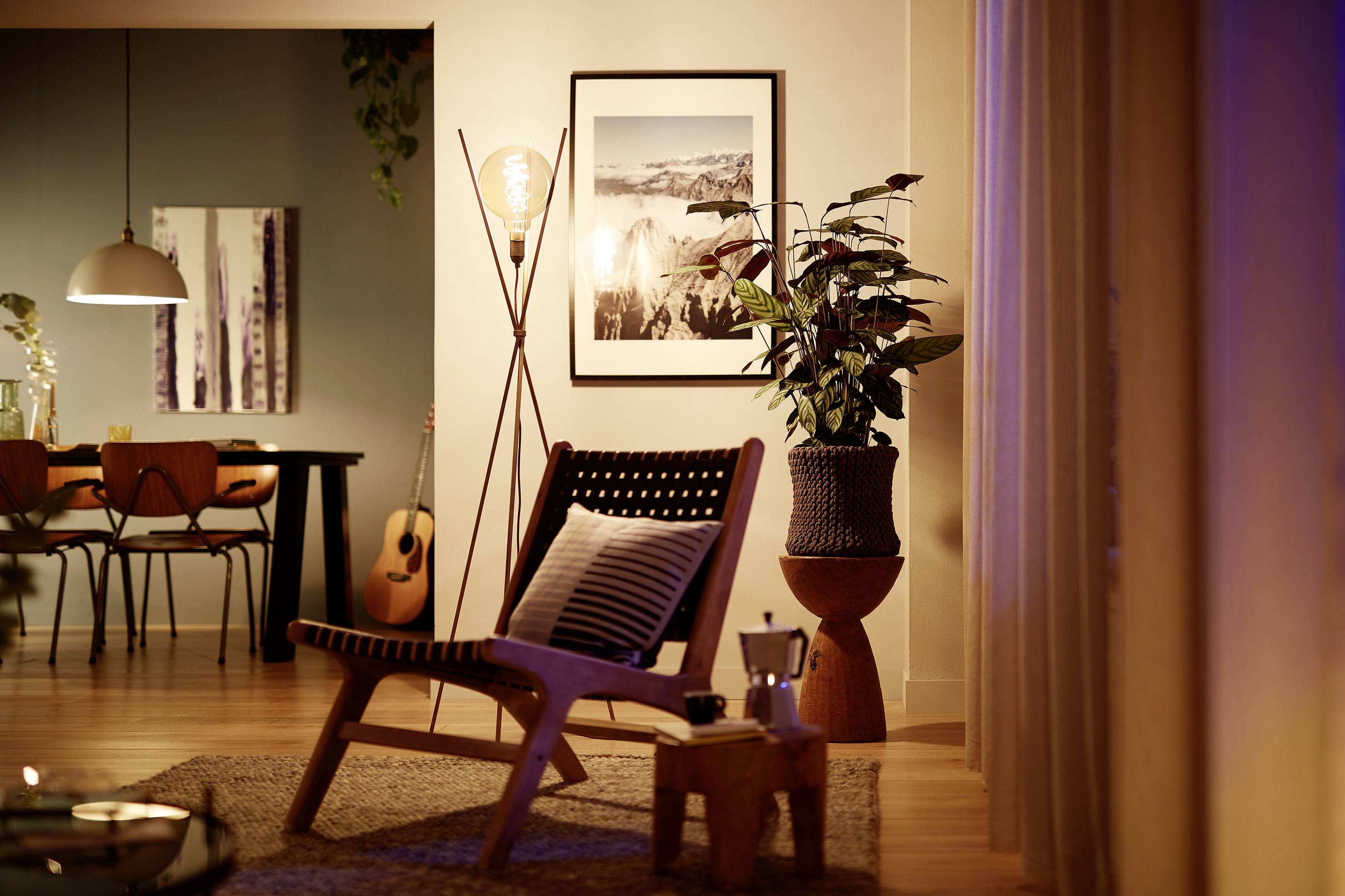 Gemütliches Wohnzimmer mit Sessel, Topfpflanze und Gitarre. Gedämpfte Beleuchtung, Wandkunst und Tisch mit Essbereich im Hintergrund.